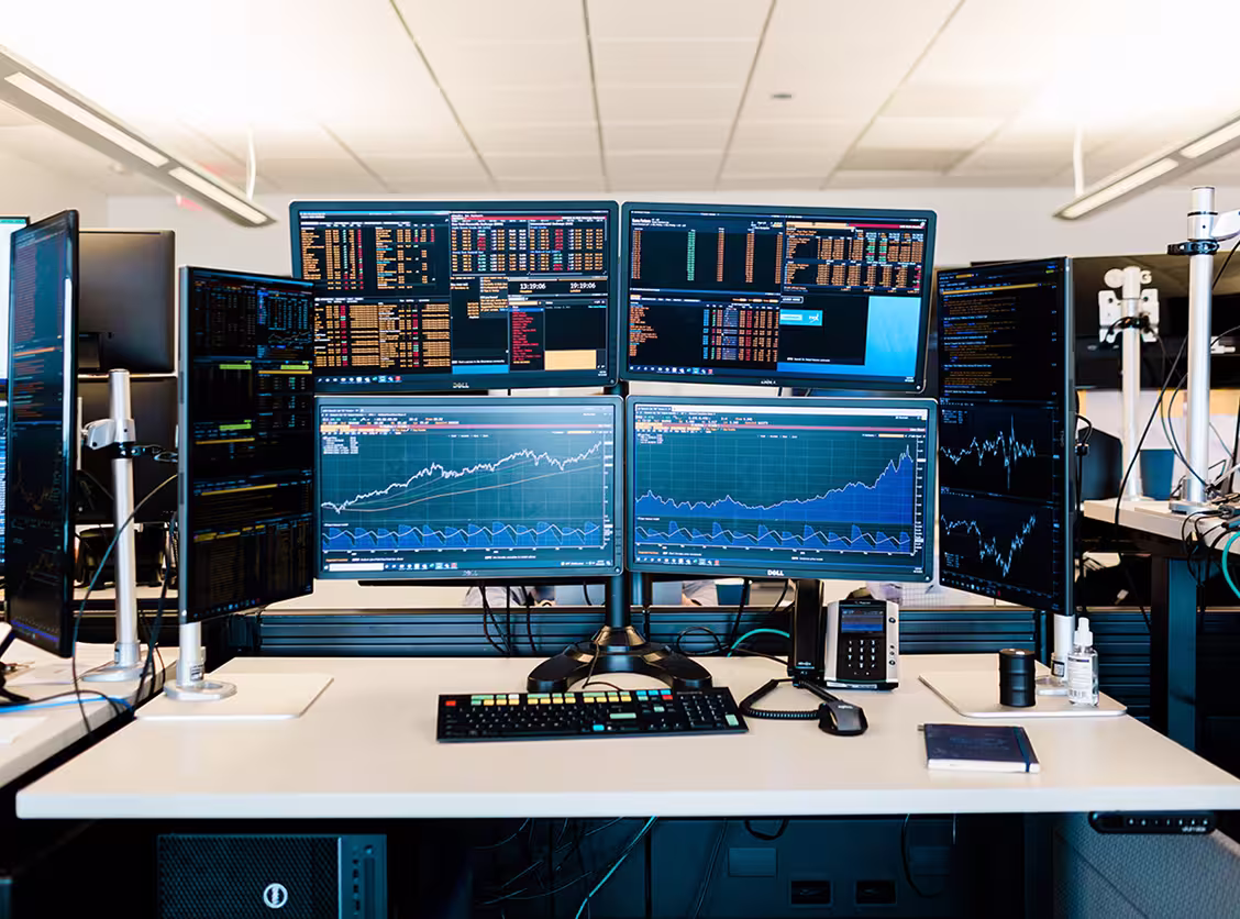 Trading desk with six monitors showing stock charts and financial data, keyboard, mouse, and phone on desk.