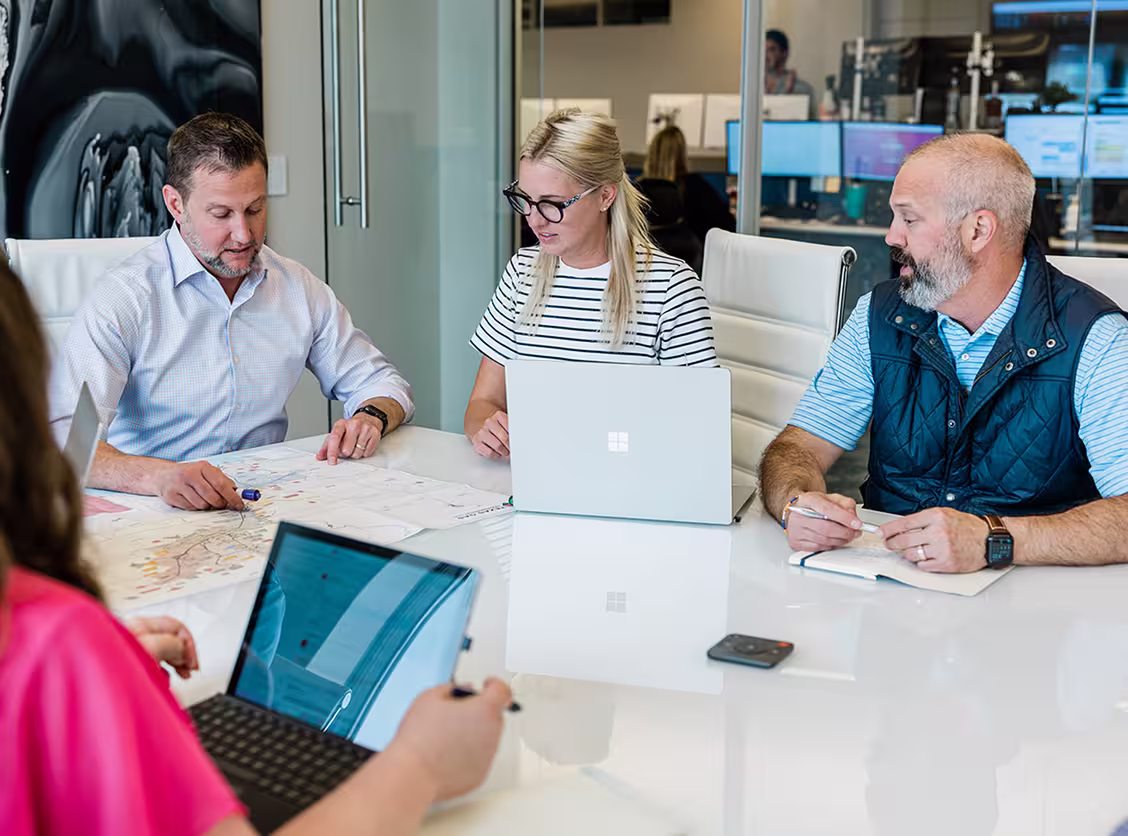 Three professionals discussing a map with two using laptops around a white conference table.