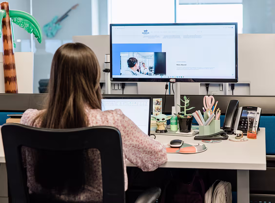 Woman with long hair working at a desk with dual monitors and office supplies in a modern workspace.