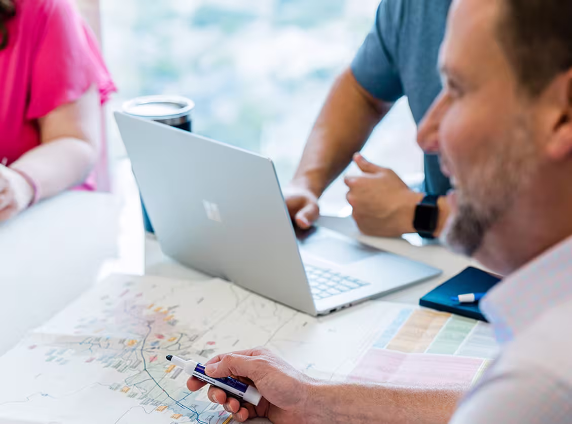 Two men and a woman in a meeting with a laptop and map on the table, one man holding a dry-erase marker.