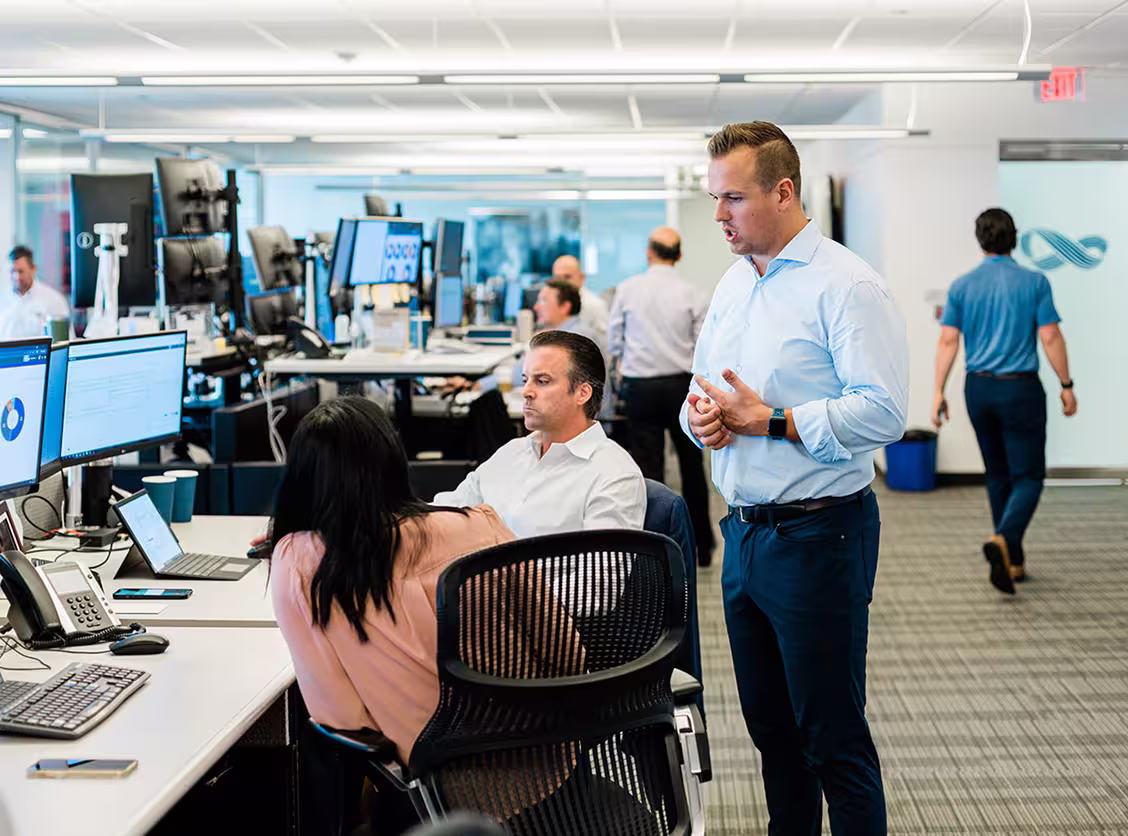 Office workspace with employees discussing near desks with multiple computer monitors and laptops.