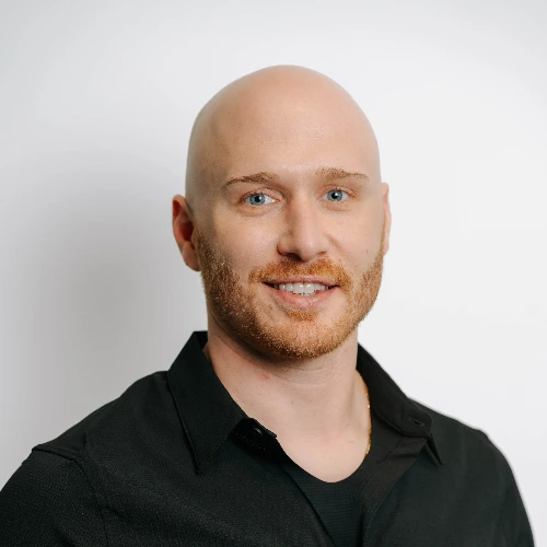 A smiling, bald man with a short reddish-brown beard wearing a black shirt against a white background.