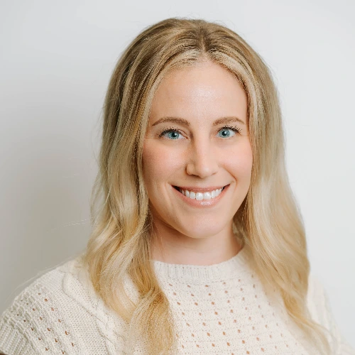 A smiling woman with wavy blonde hair wearing a sleeveless cream knit sweater against a light background.