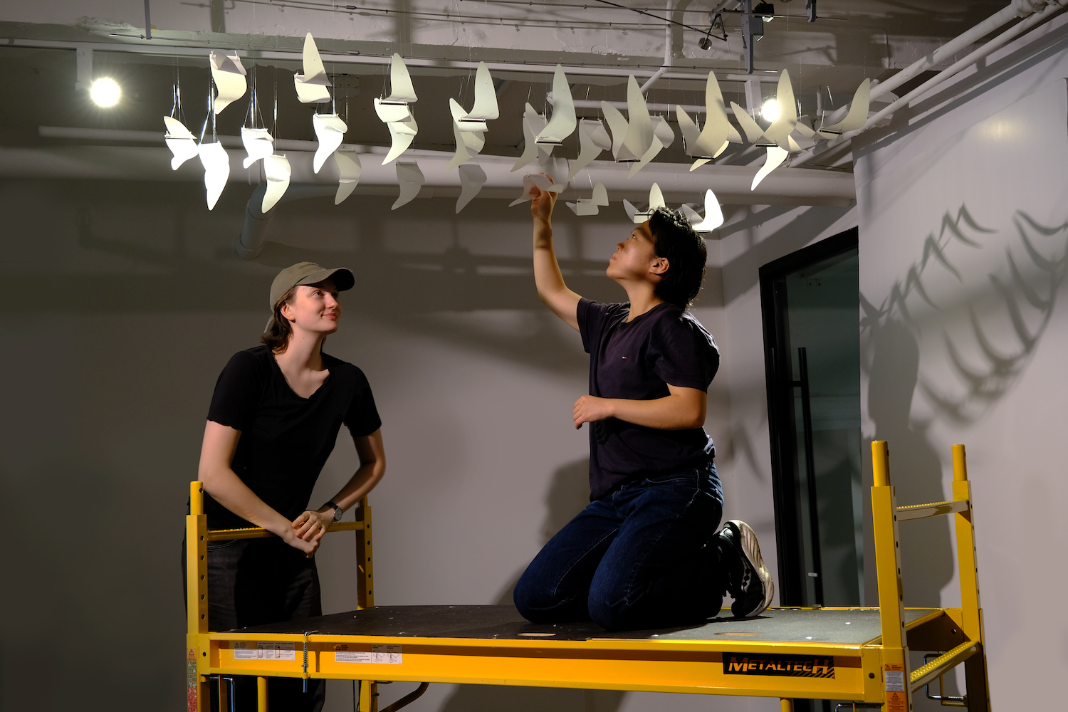 Photo of Pepi Ng and me sitting on yellow scaffolding, installing birds to the sculpture on the ceiling. 