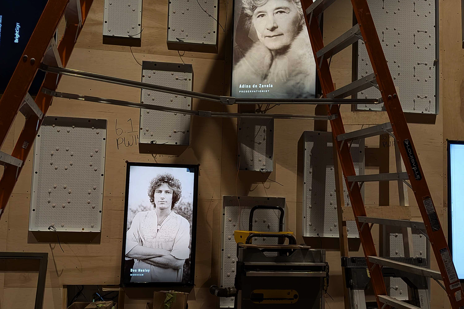 Installation setup with two portrait screens surrounded by empty mounting panels and a ladder.