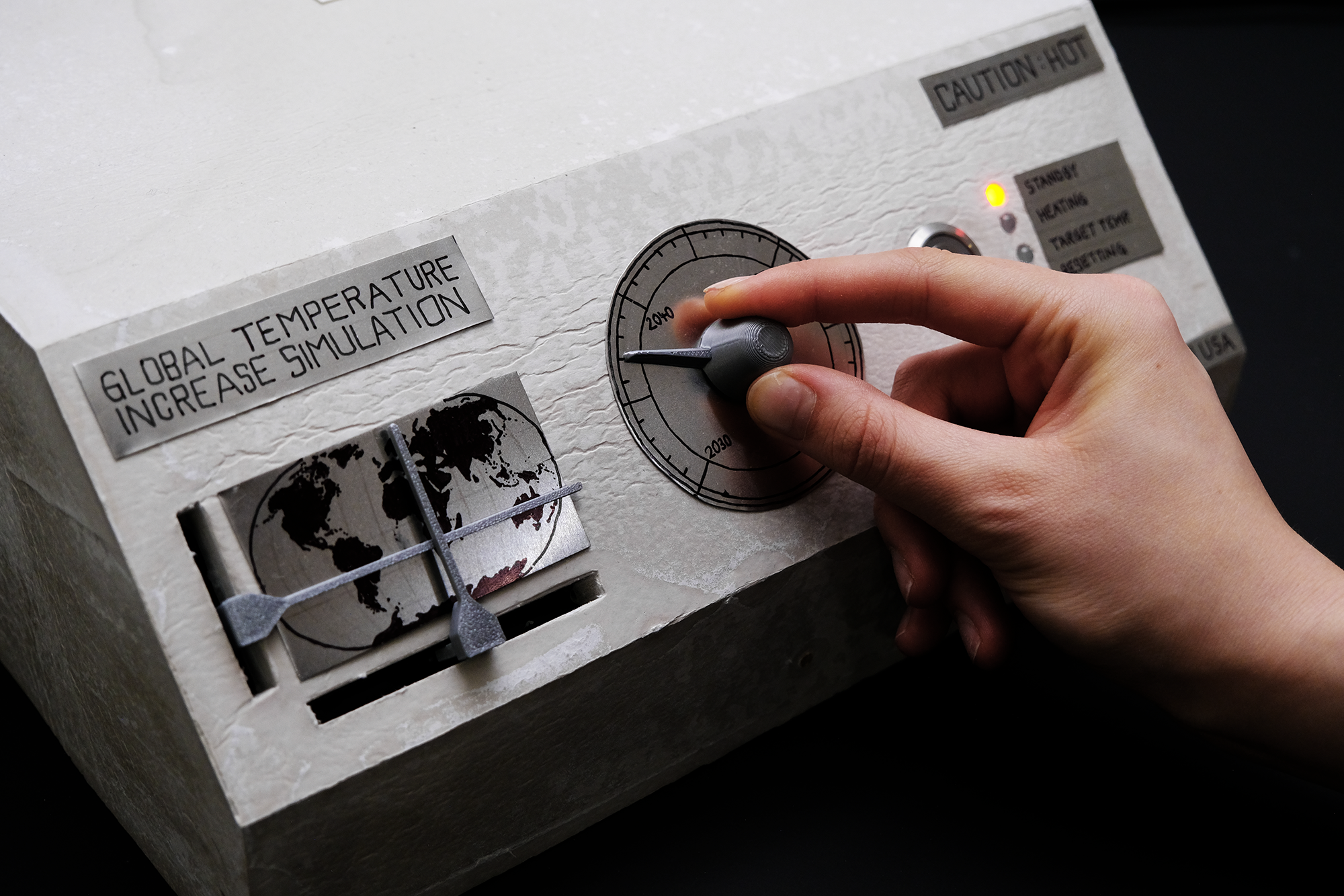 Hand adjusting a dial on a device labeled 'Global Temperature Increase Simulation' with a world map graphic and a caution sign for heat.