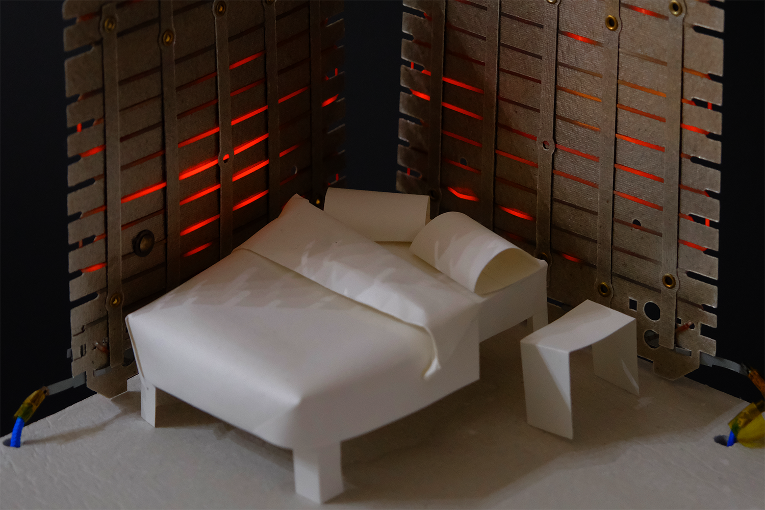 Miniature paper model of a bed with pillows and a side table, set against a backdrop with glowing red light.
