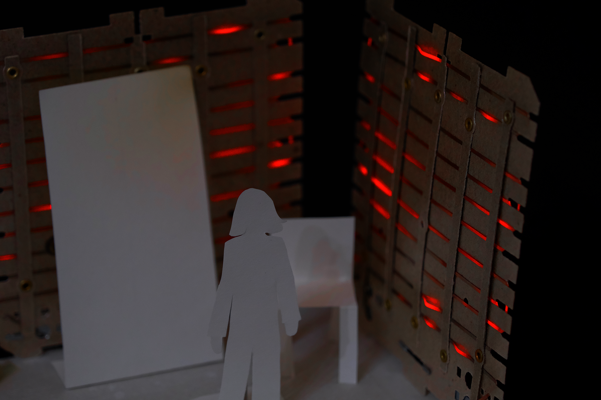 Paper cutout figure standing in front of paper furniture and a cardboard backdrop lit with red light.