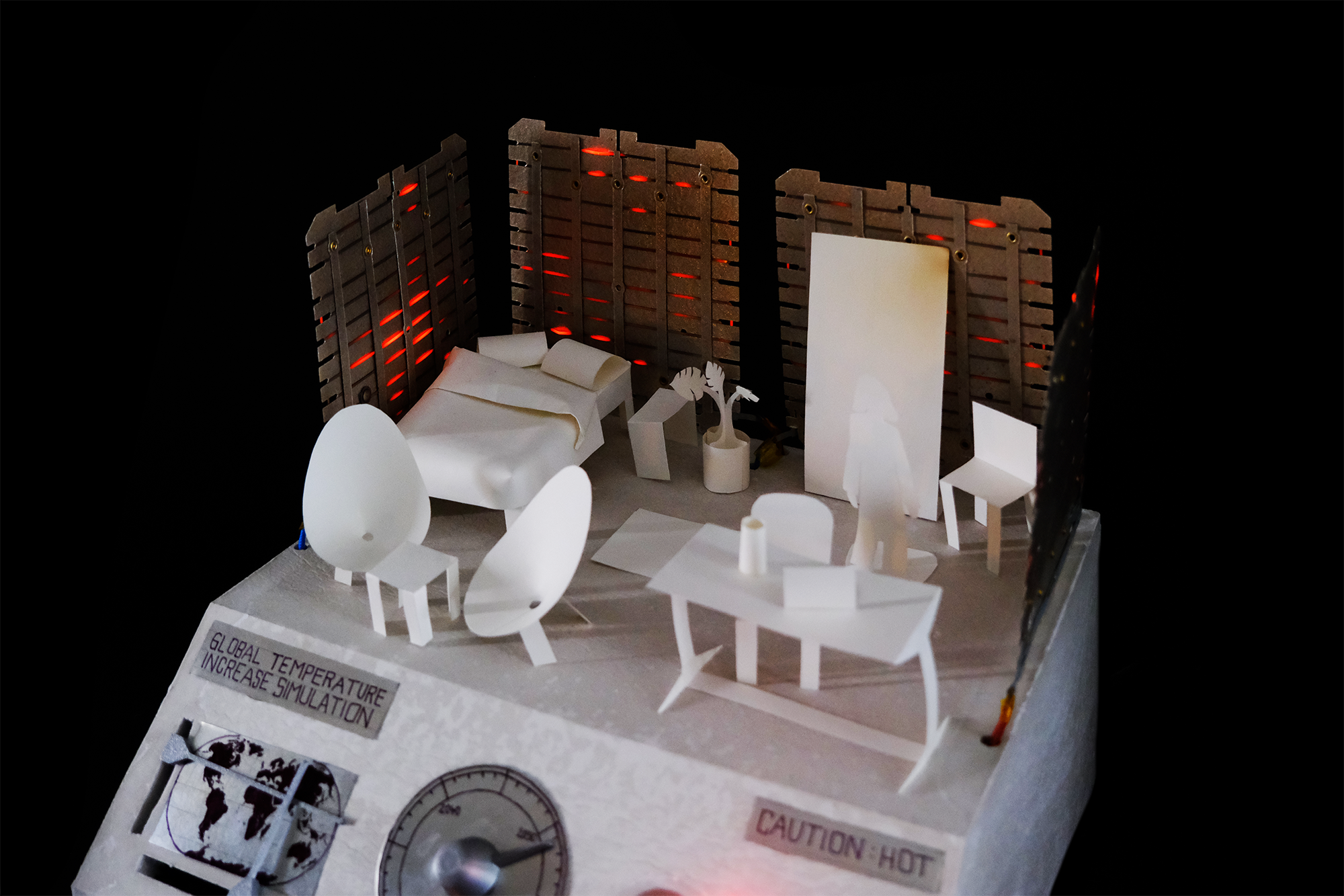 Miniature white paper model of a heated room with furniture and a figure, surrounded by dark walls with red glowing wires, labeled 'Global Temperature Increase Simulation' .