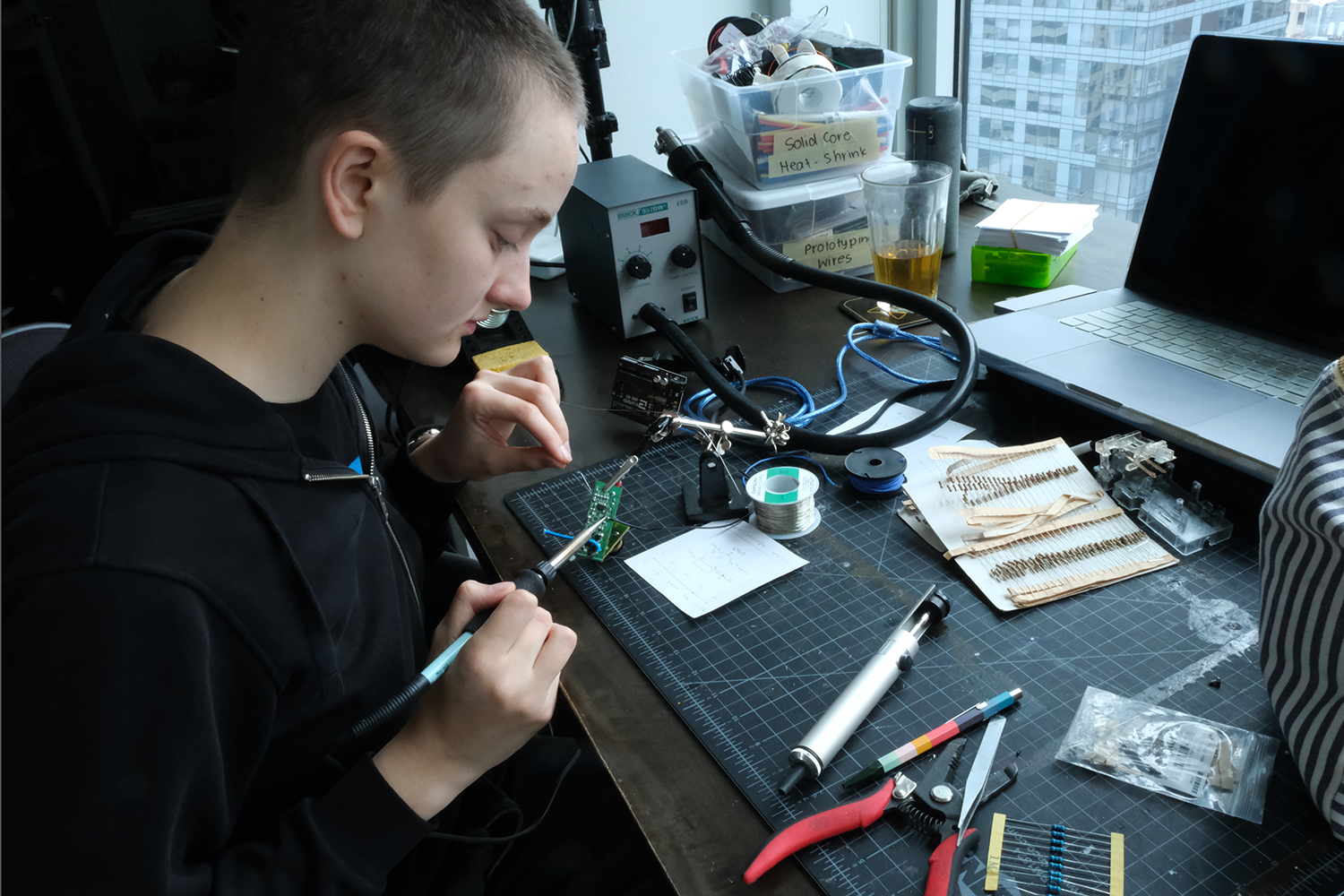 Person soldering electronic components on a circuit board at a cluttered desk with tools and a laptop nearby.