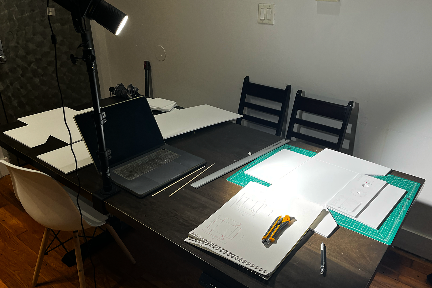 Worktable with laptop, cutting mat, sketches, utility knives, metal ruler, and a desk lamp illuminating the workspace.