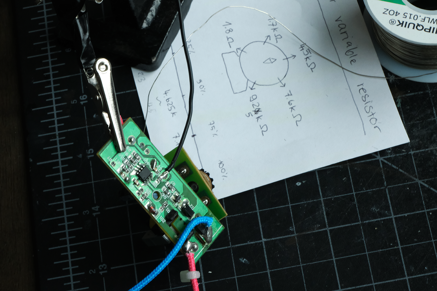 Close-up of a circuit board with wires clipped by an alligator clip, resting on a cutting mat next to a hand-drawn resistor circuit diagram and a spool of solder wire.
