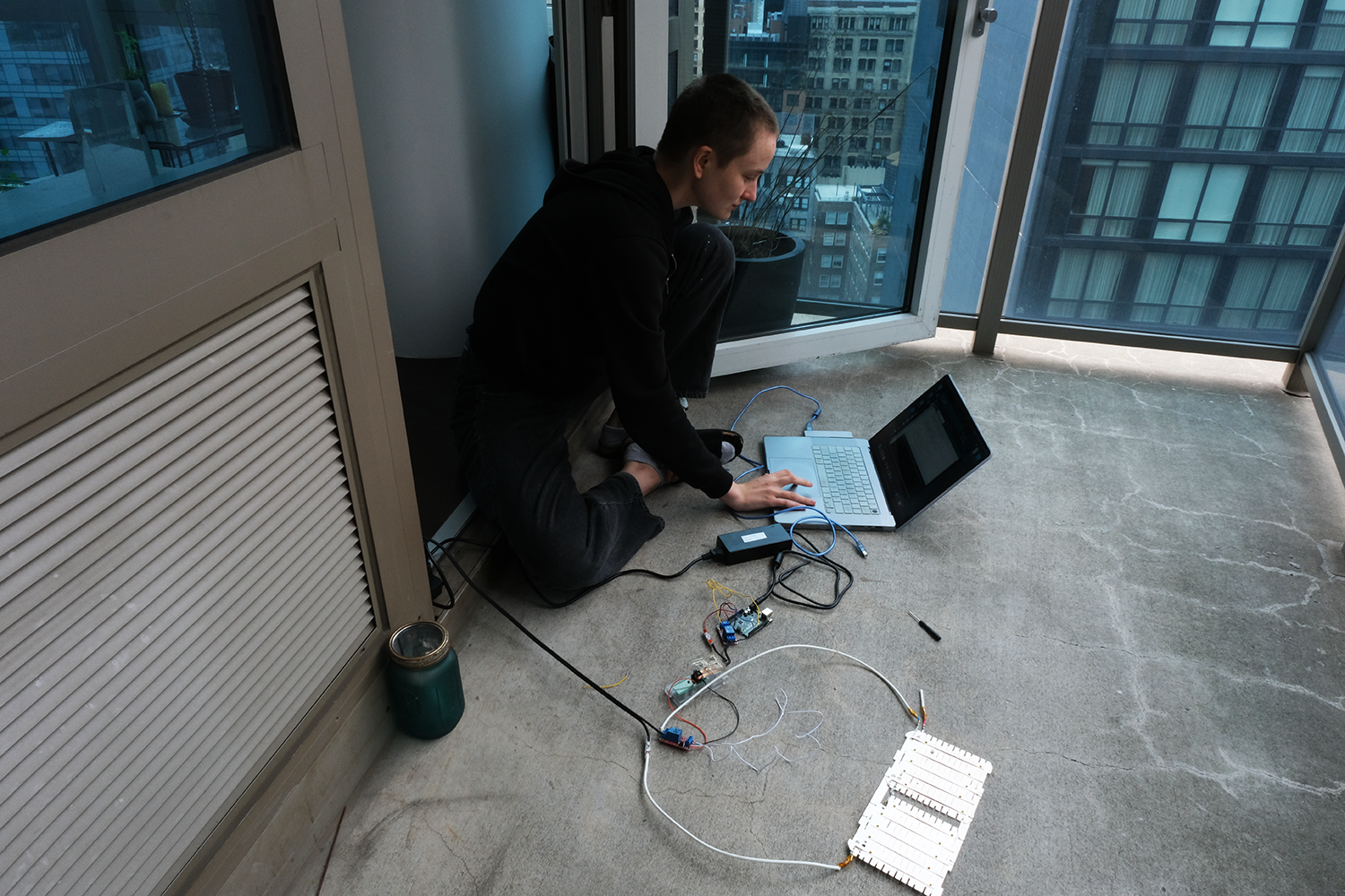 Person sitting on a balcony floor working on a laptop connected to electronic circuitry and components.