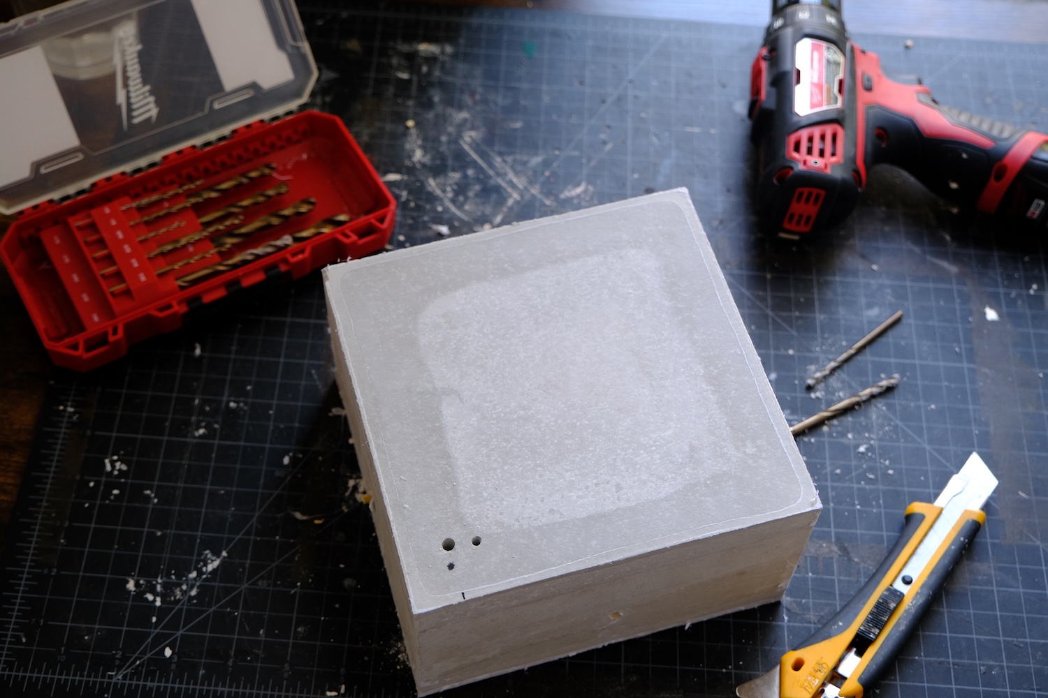Close-up of a square plaster block with drilled holes on a cutting mat, surrounded by drill bits in a case, a cordless drill, and a utility knife.
