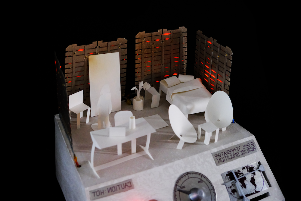 Miniature white model of a room with bed, chairs, table, plant, and figure, surrounded by dark walls with glowing red light and a temperature dial labeled 'Caution: Hot' and 'Global Temperature Increase Simulation'.
