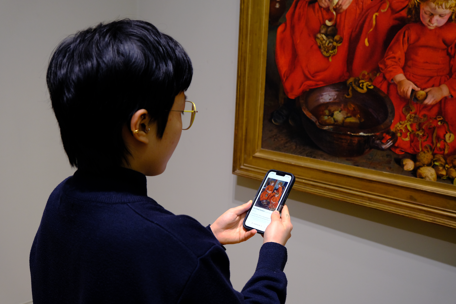Person with short dark hair and glasses holding a smartphone displaying information about a painting, standing near the same painting of children peeling potatoes in red dresses.
