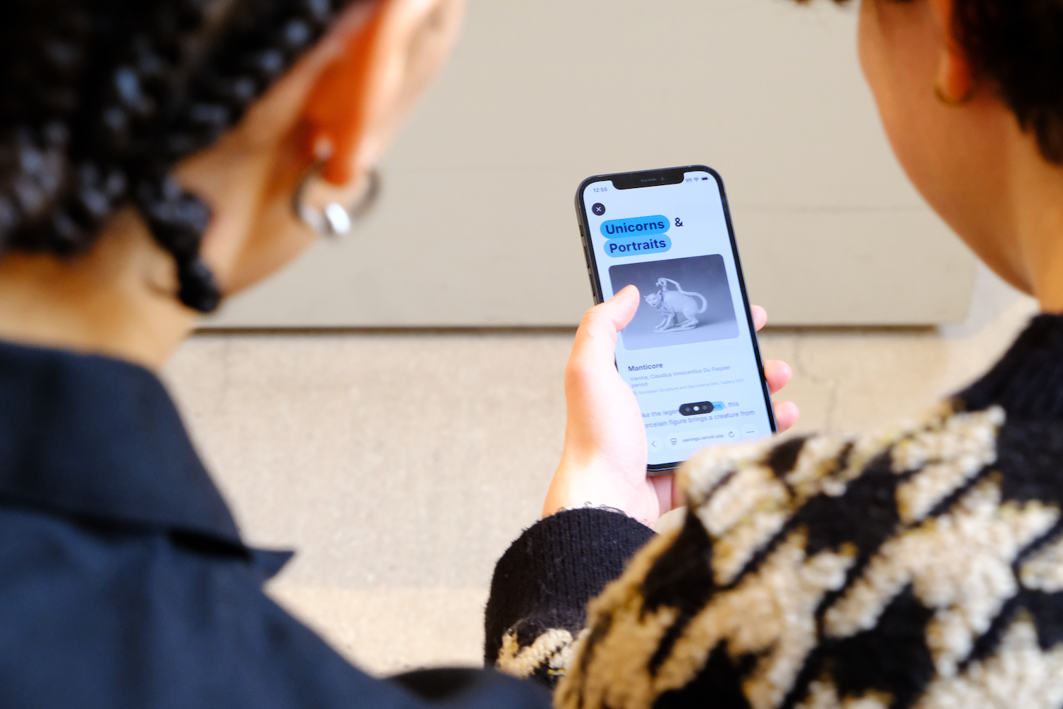 Two people viewed from behind looking at a mobile phone screen displaying a page titled 'Unicorns & Portraits' with an image of a mythical creature sculpture.