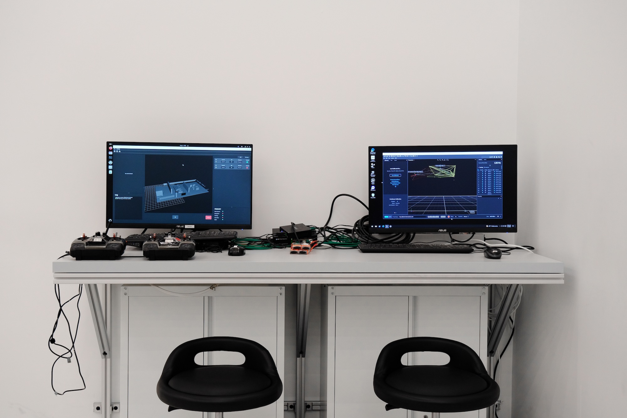 Two computer monitors on a table showing 3D modeling software, with two black chairs in front.