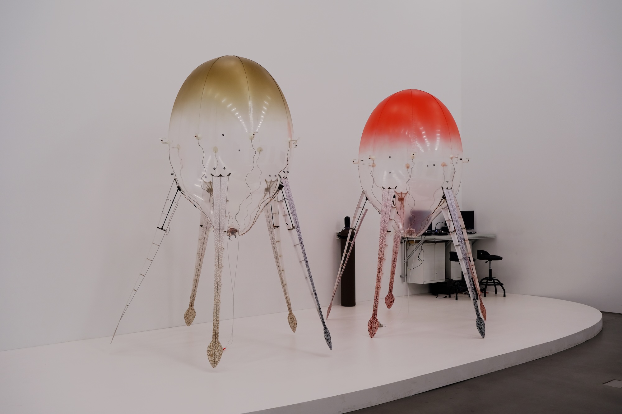 Two large translucent balloon-like art sculptures with long legs, one gold-tinted and one red-tinted.
