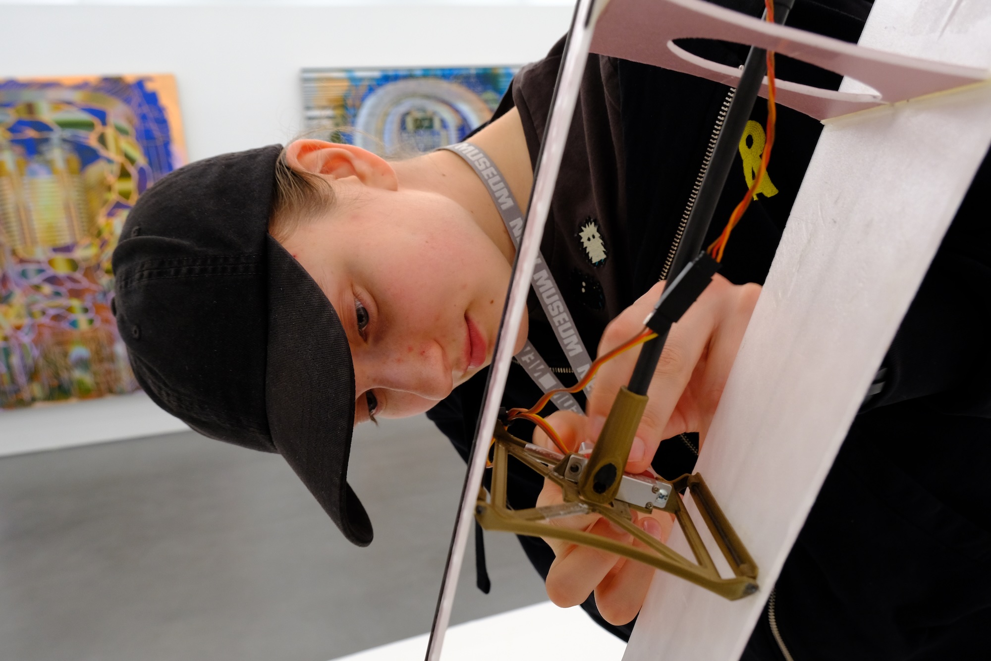 Person in black cap working with a mechanical device on a white table in an art gallery.
