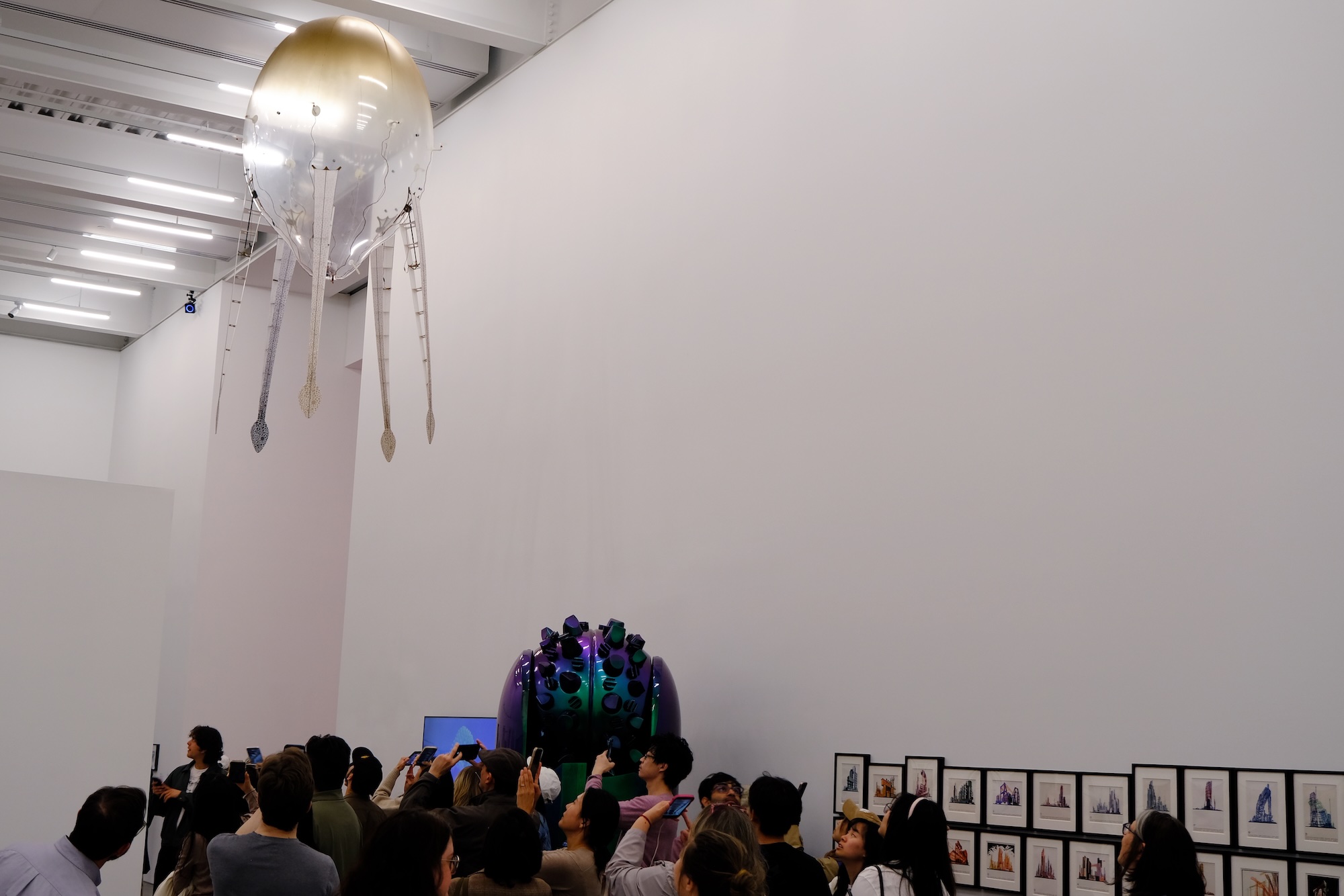 Group of people in an art gallery viewing a large hanging balloon-shaped sculpture and framed artworks on the wall.