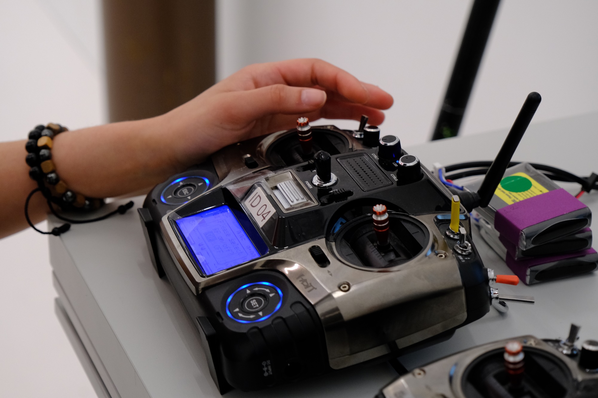 Hand resting over joystick on radio controller device with antennas and display on table.