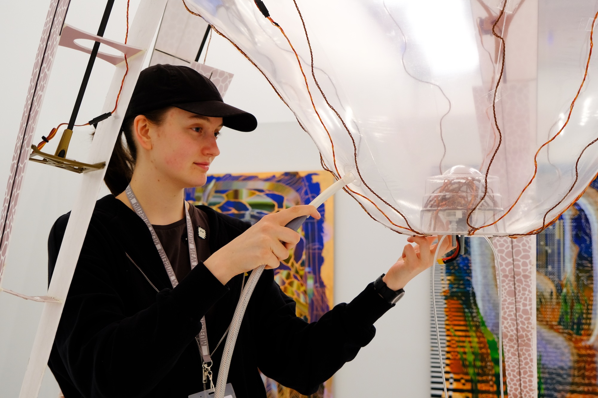 Person in black cap inflating a large transparent balloon-like structure with wires.
