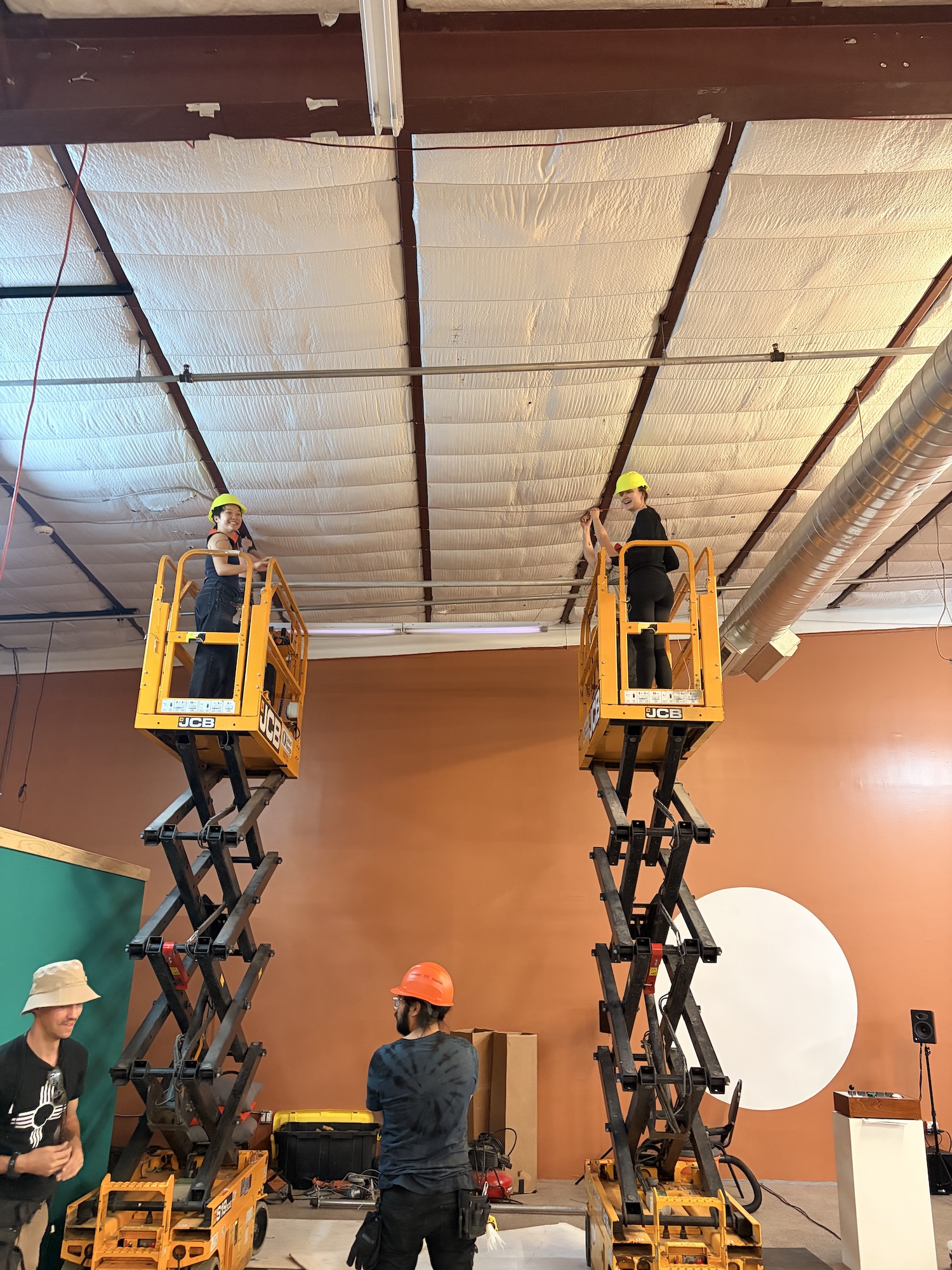 Two people on yellow forklifts working near ceiling with two others wearing helmets on ground.