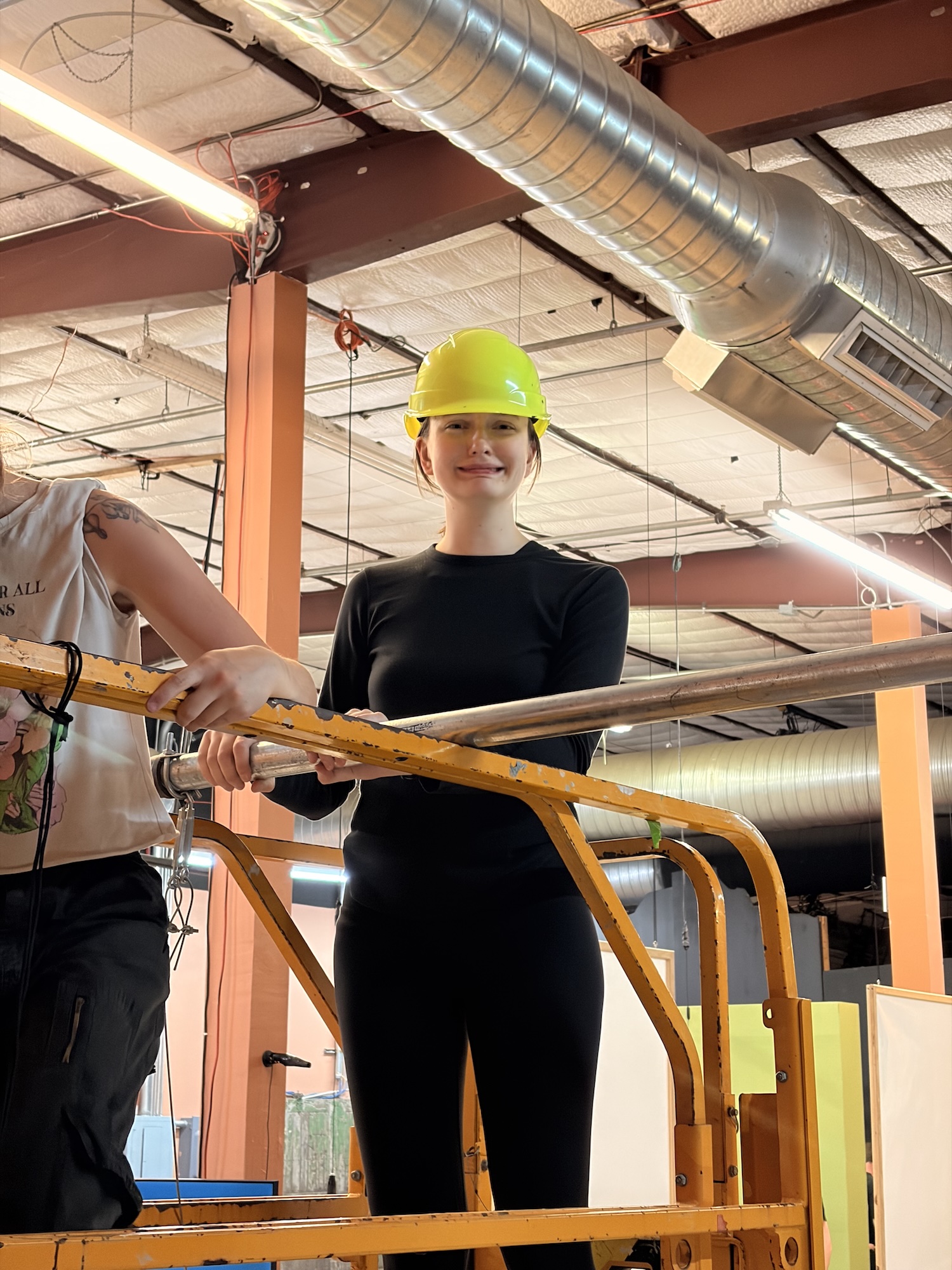 Person in black outfit wearing a yellow safety helmet inside industrial facility near orange railing.
