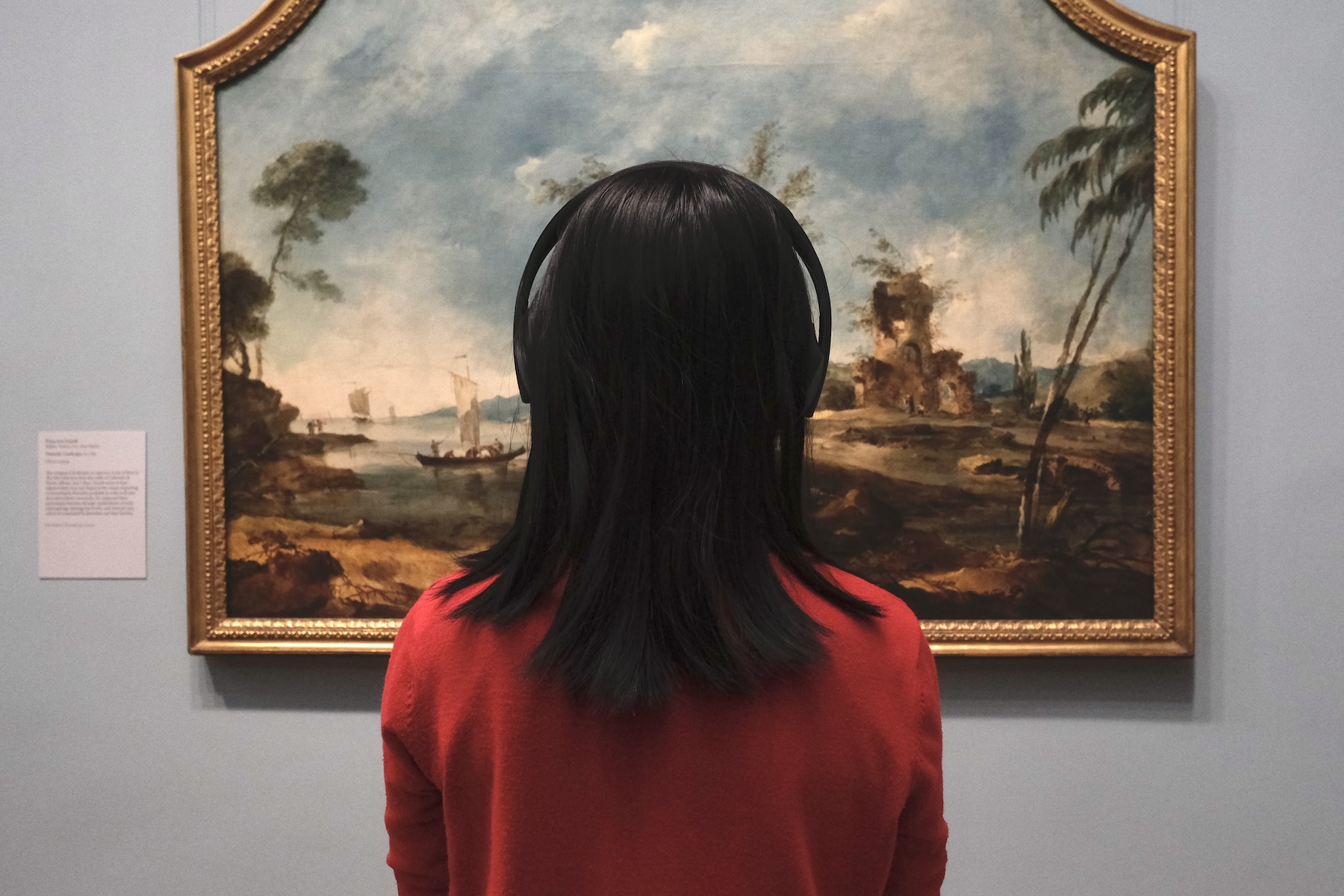 Person in red sweater and headphones views a framed landscape painting with boats and ruins.