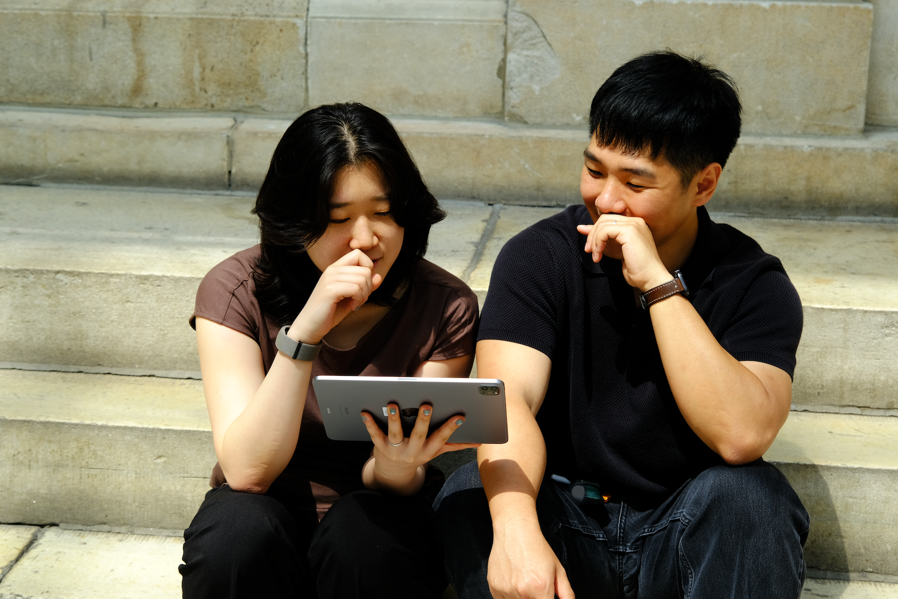 Two people sitting on stone steps, looking at a tablet and smiling thoughtfully.