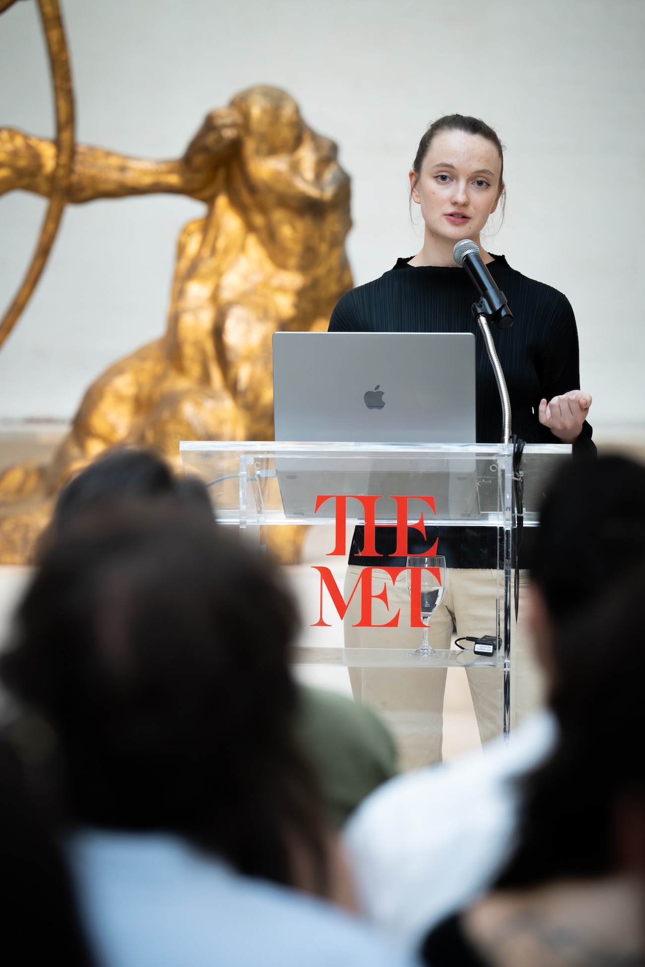 Woman speaking at The Met podium with laptop and microphone to an audience in a gallery.