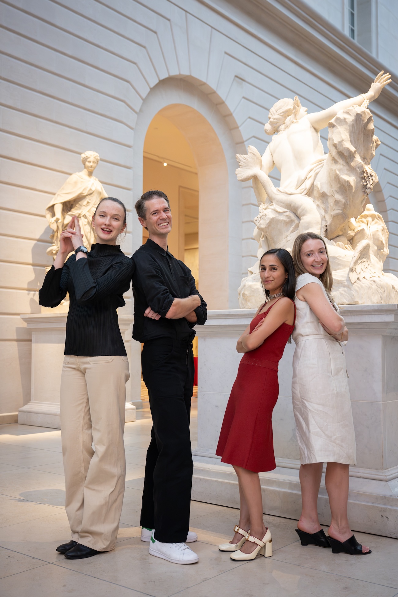 Four people posing with arms crossed and smiling inside a museum with classical marble sculptures.