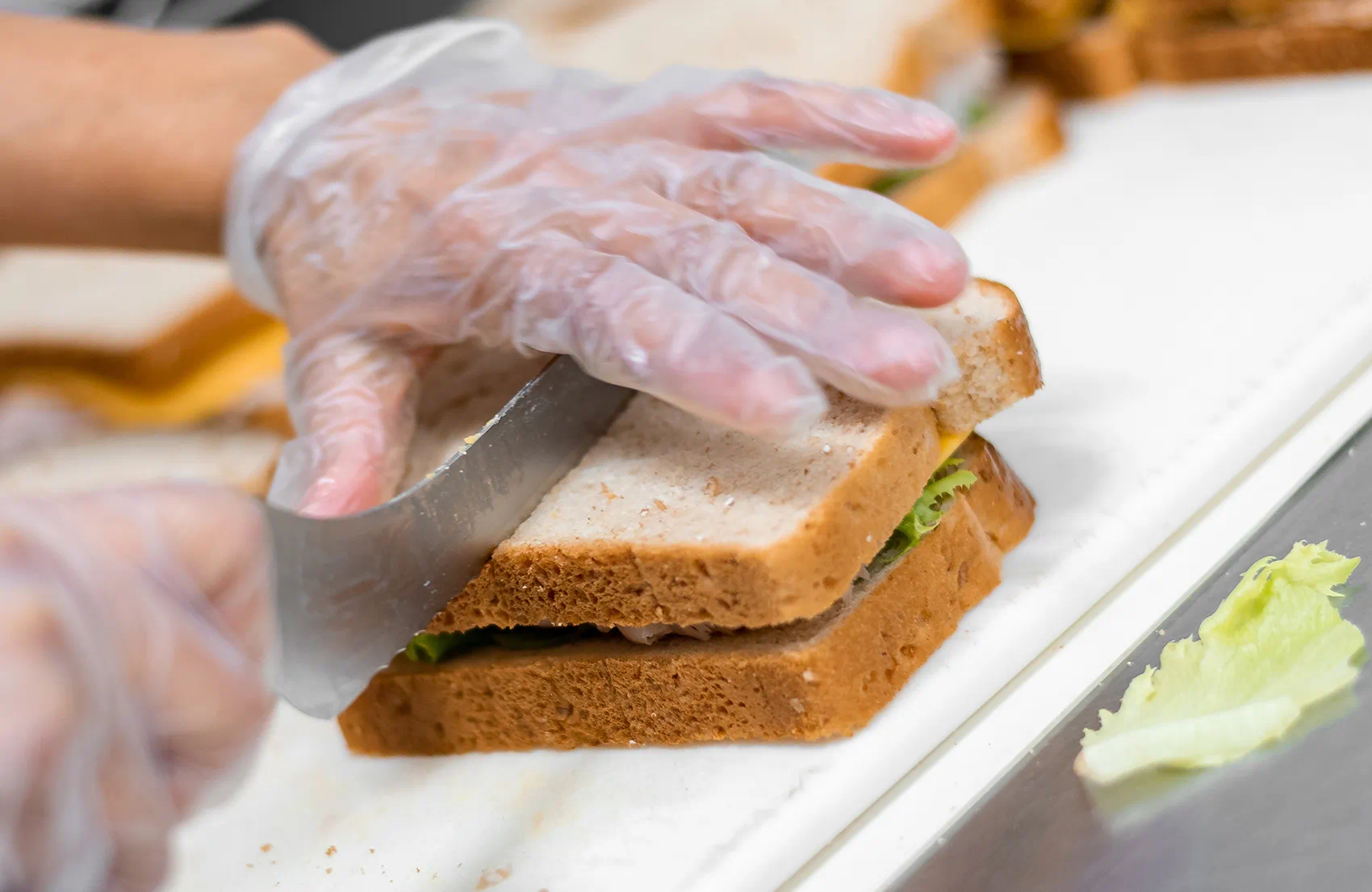 Person wearing gloves slicing a sandwich with lettuce on a white cutting board.