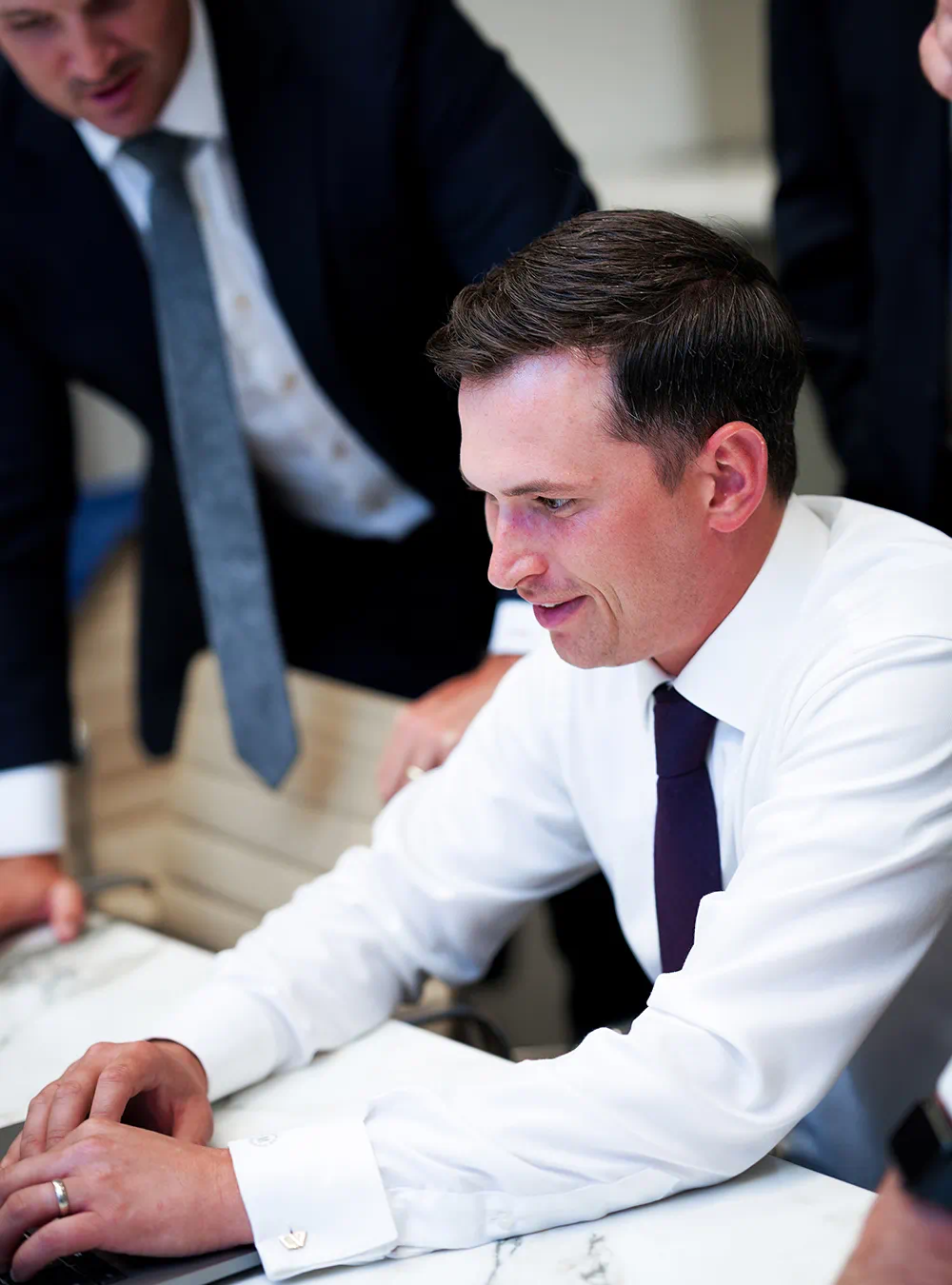 Man in white shirt and dark tie working on a laptop with colleagues in business attire around him.