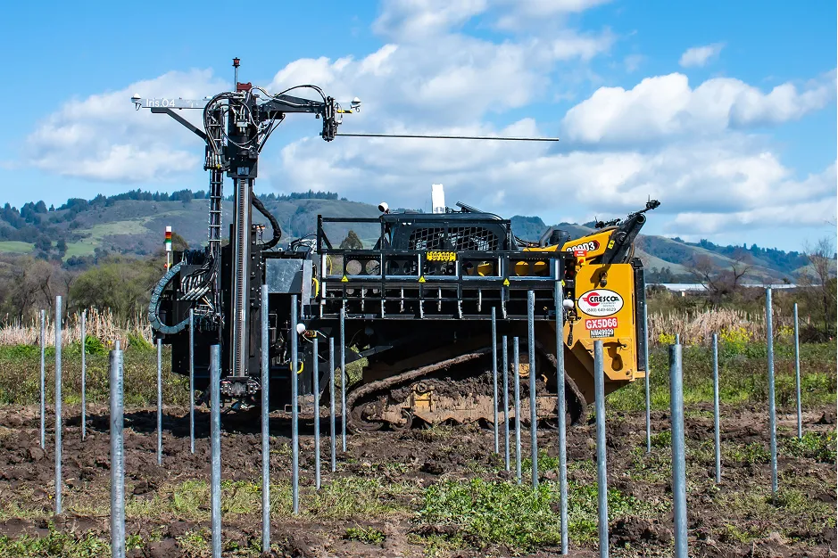 Tracked machine installing metal posts for a solar array on a muddy field with hills in the background