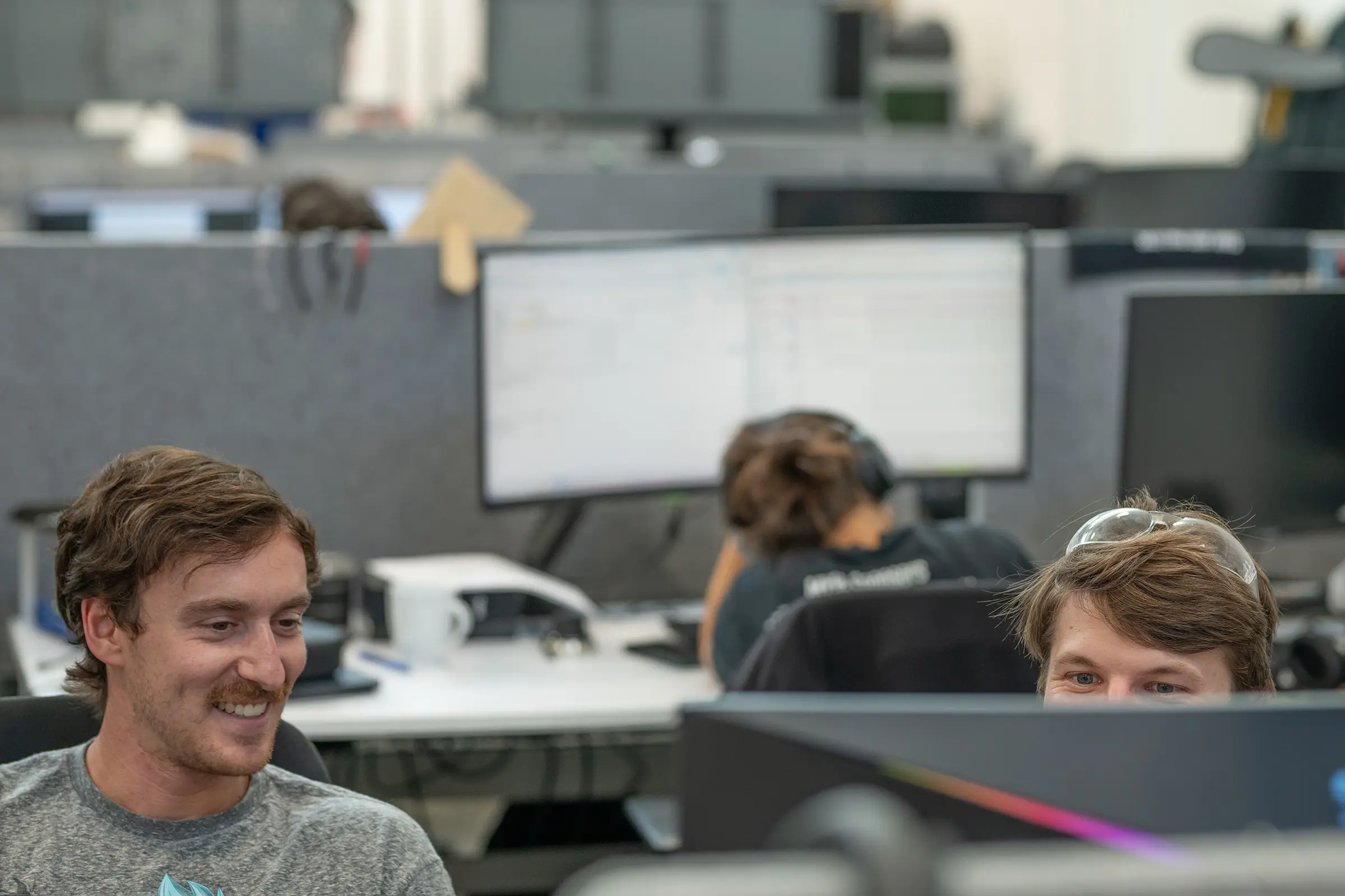 Two coworkers smile and look at a computer monitor in an open-plan office, with another person working at a desk in the background.
