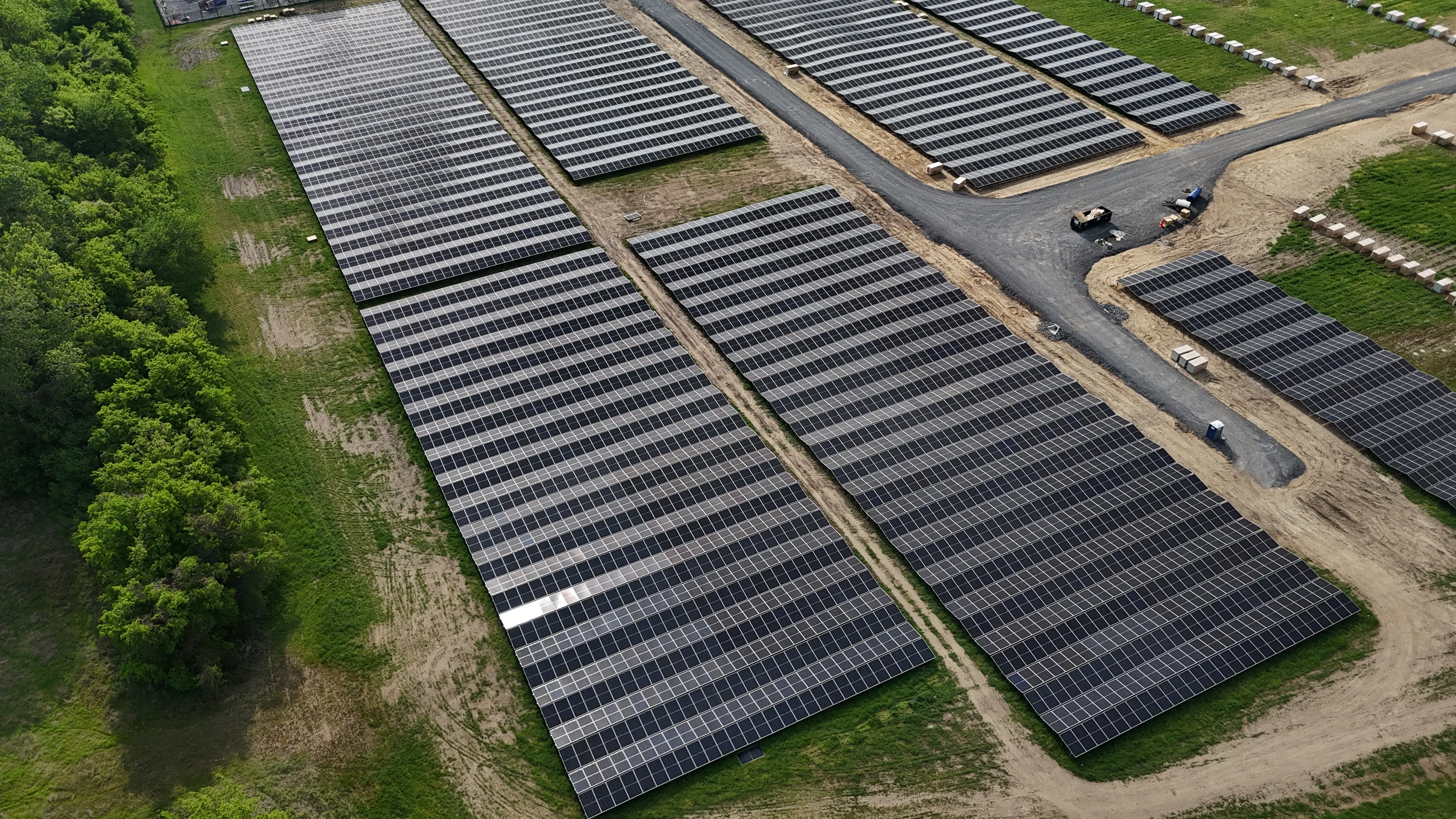 Aerial view of a large solar farm with multiple rows of solar panels arranged in rectangular sections on green grass and dirt paths, adjacent to a wooded area with dense green trees.