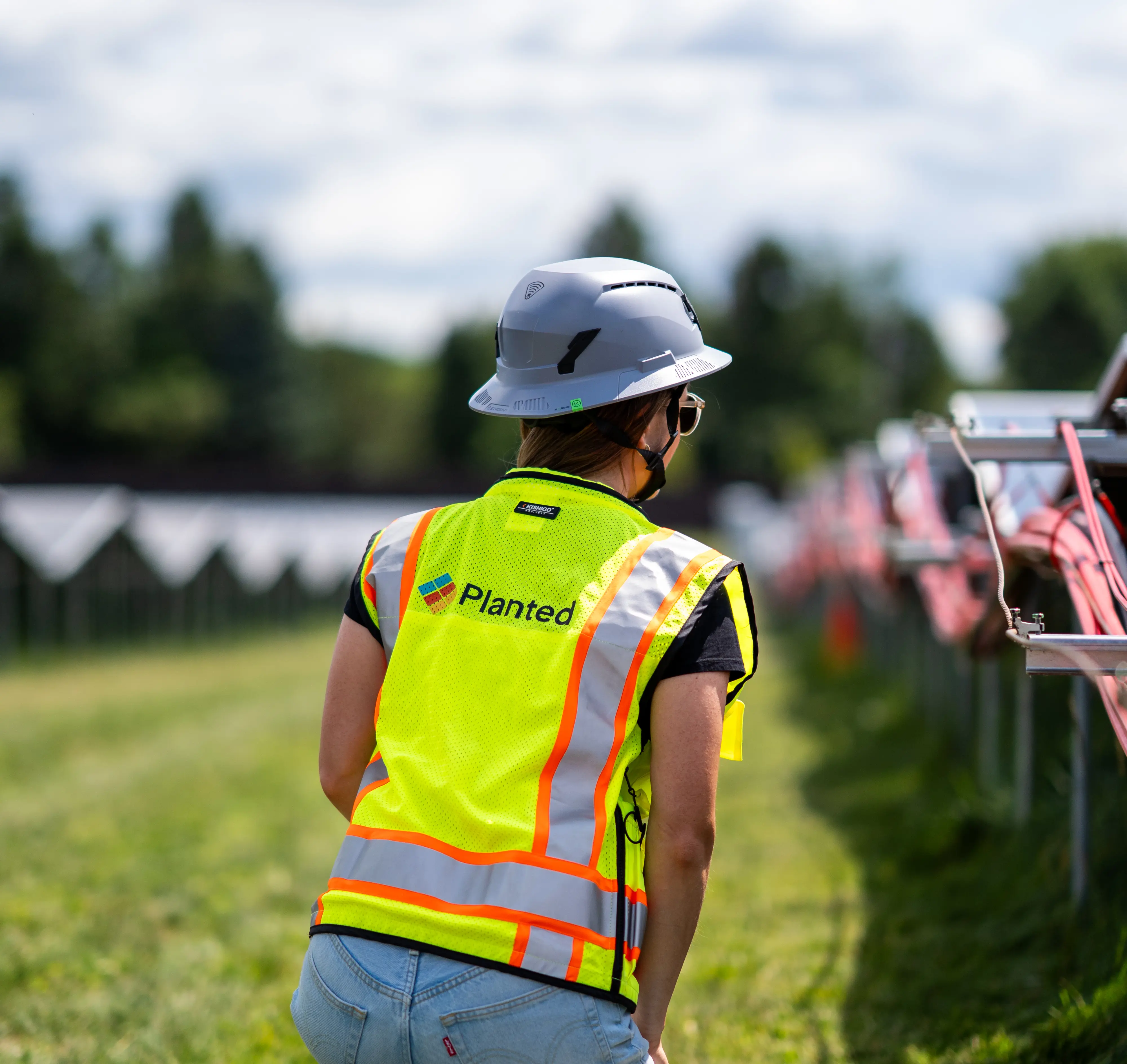 A planted solar worker wearing a gray hard hat and bright yellow reflective safety vest stands beside a row of solar panels in a grassy field.
