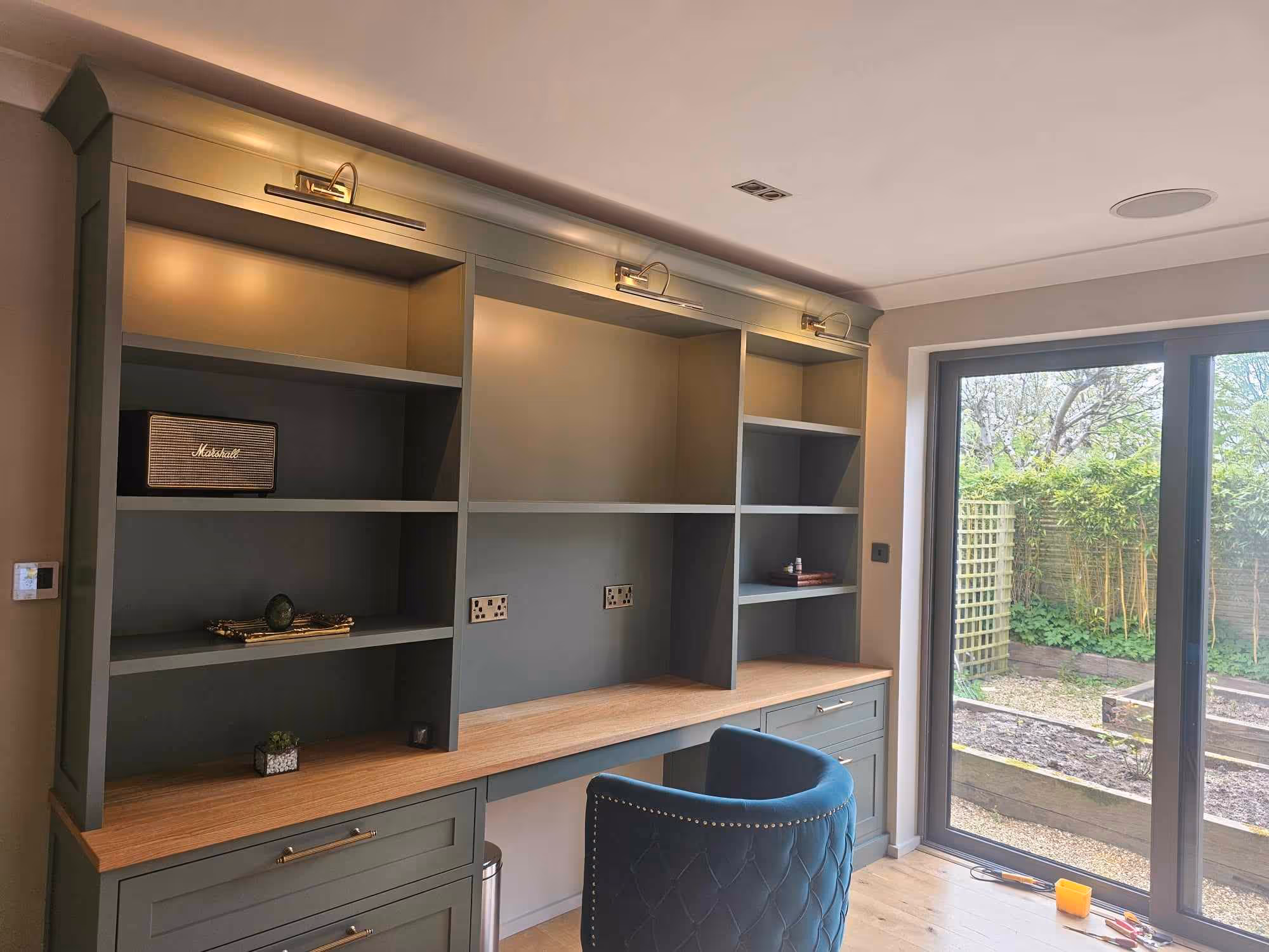 Modern home office with green built-in shelving and wooden desk, blue quilted chair, and glass sliding door overlooking garden.