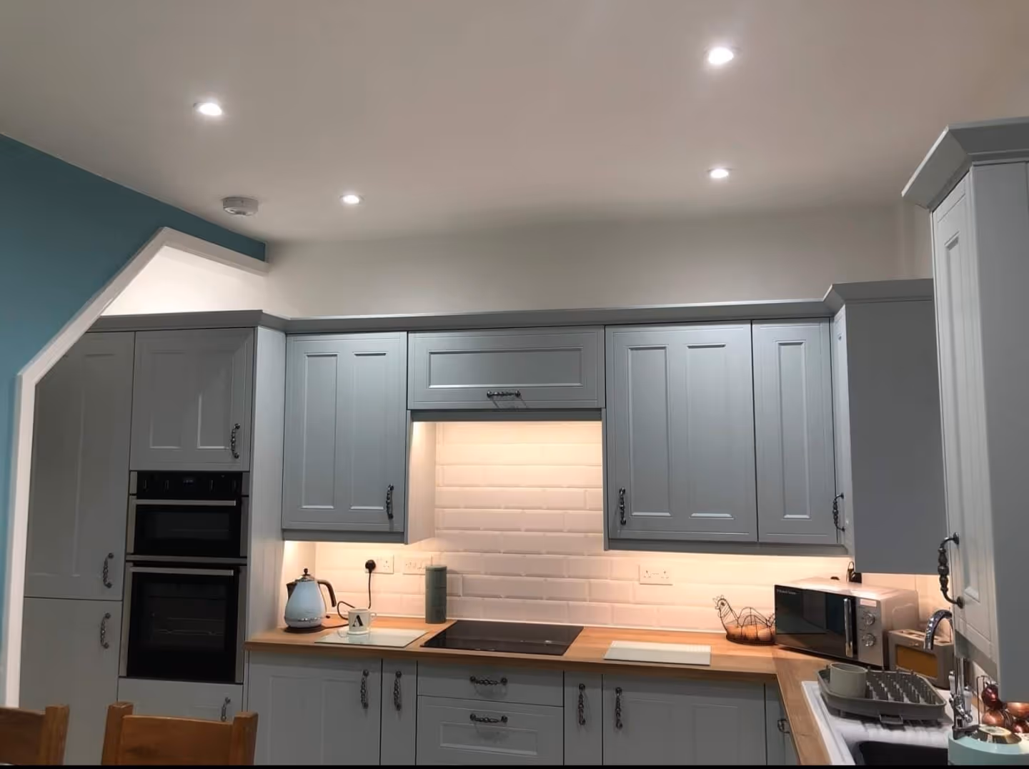 Modern kitchen with grey cabinets, wooden countertop, built-in oven, electric hob, kettle, microwave, and sink with dish rack.