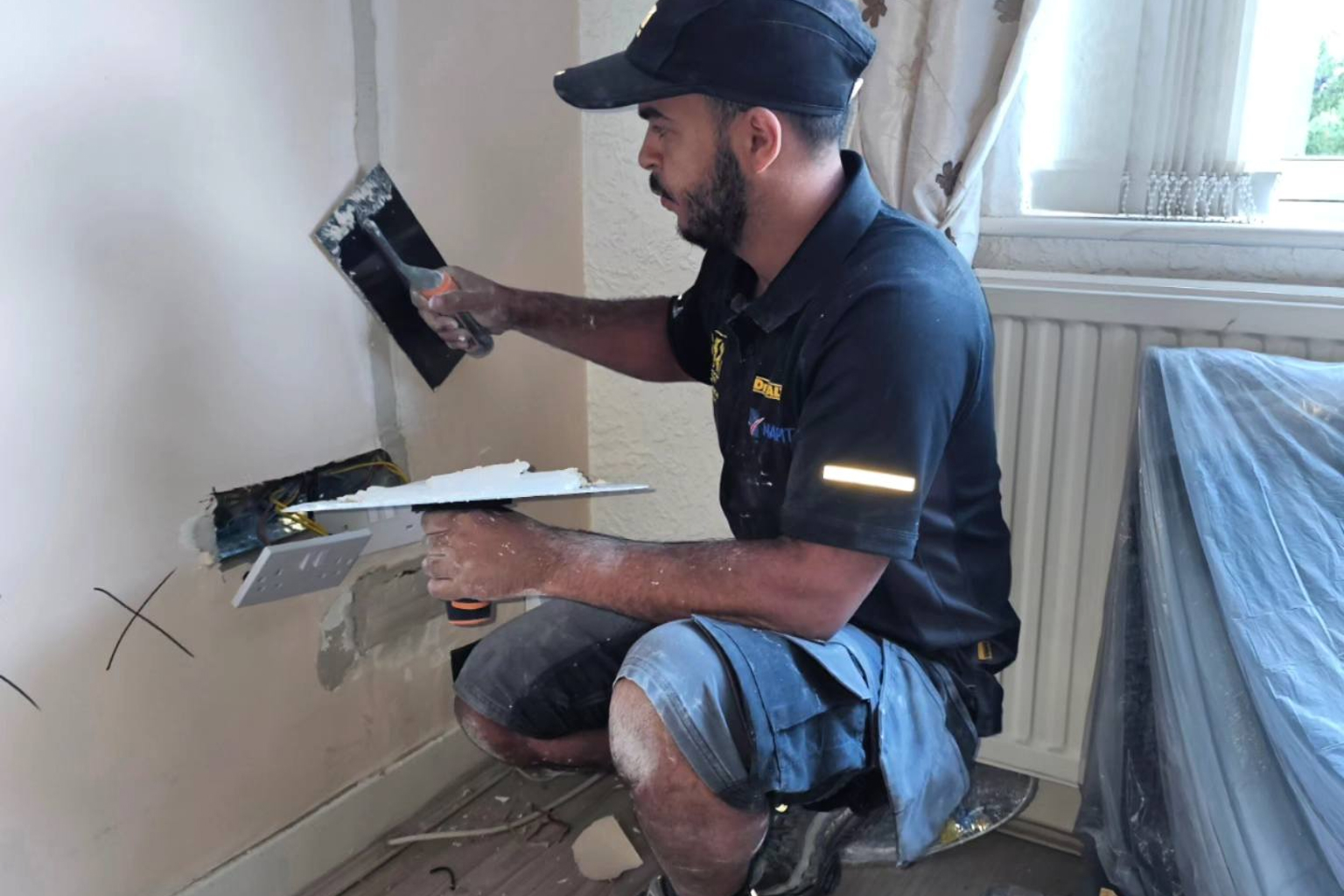 Electrician kneeling while plastering around a wall socket with exposed wiring.