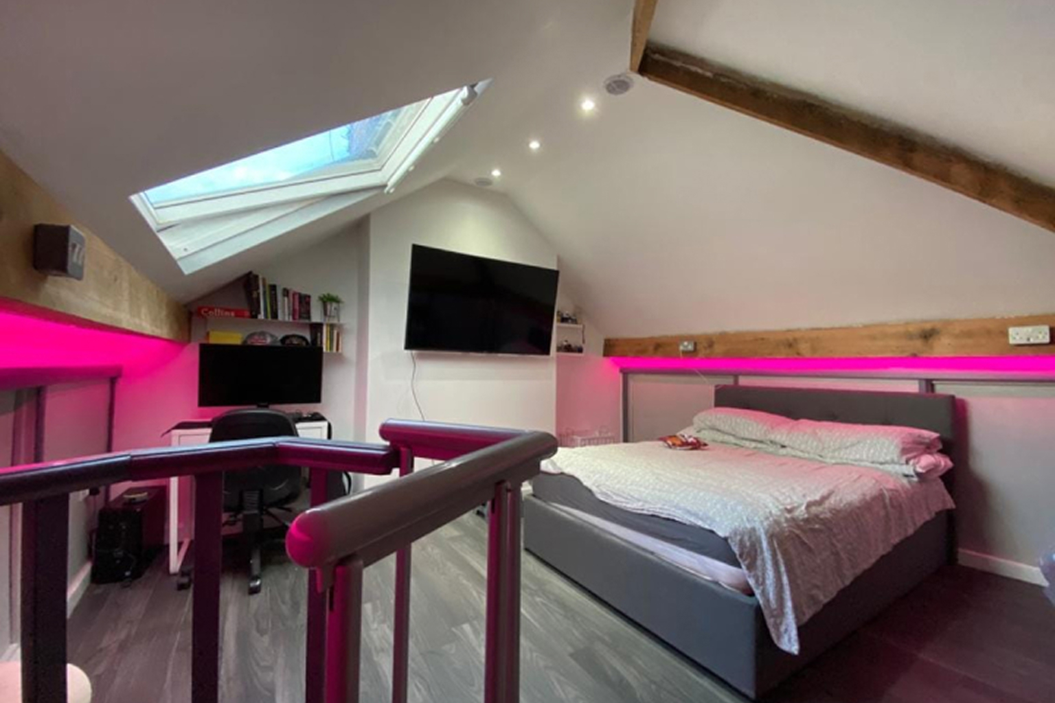 Modern attic bedroom with grey upholstered bed, skylight window, wall-mounted TV, desk with computer, and pink LED accent lighting under wooden beams.