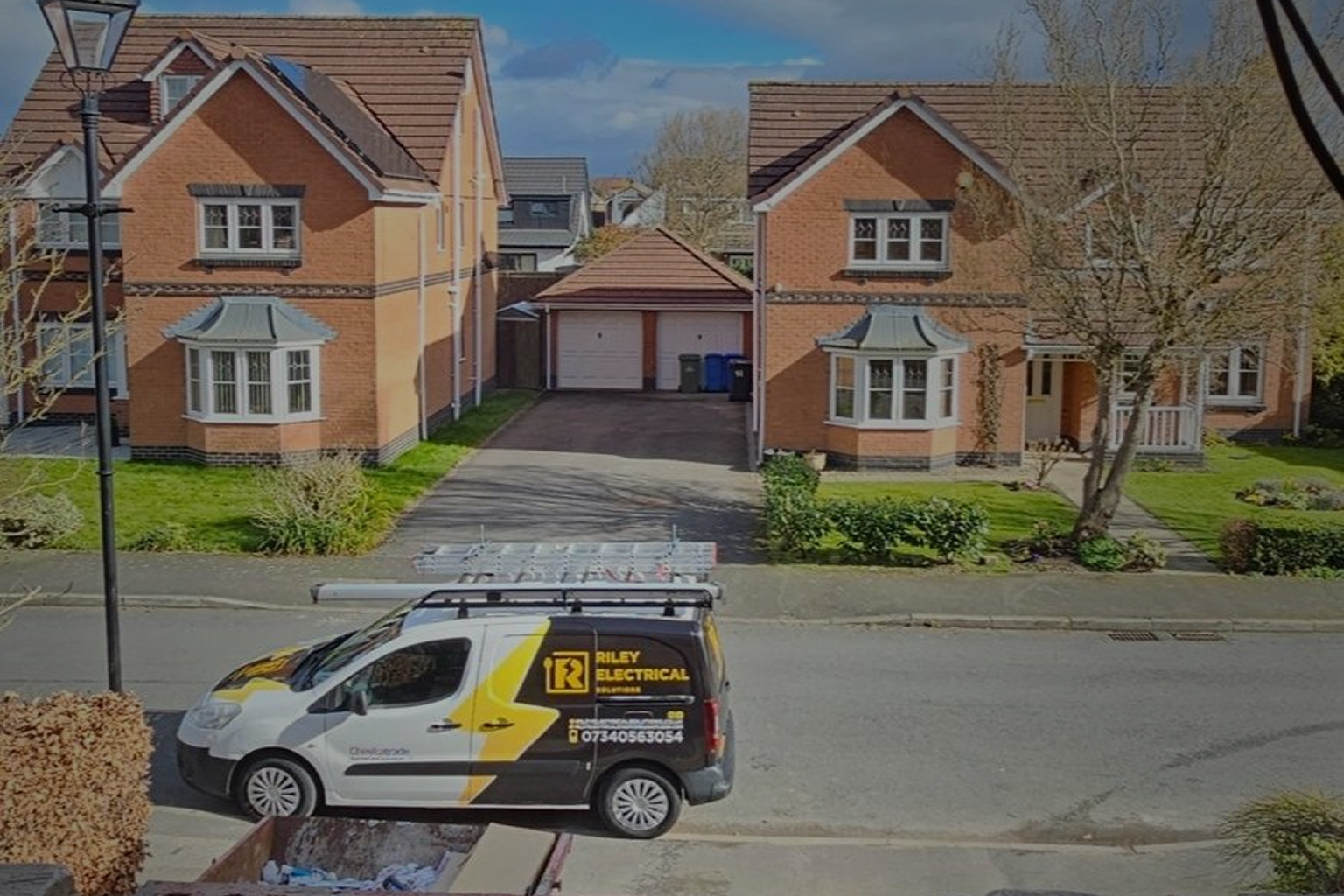 White and black Riley Electrical Solutions van parked on a residential street in front of two brick houses with bay windows and green lawns.