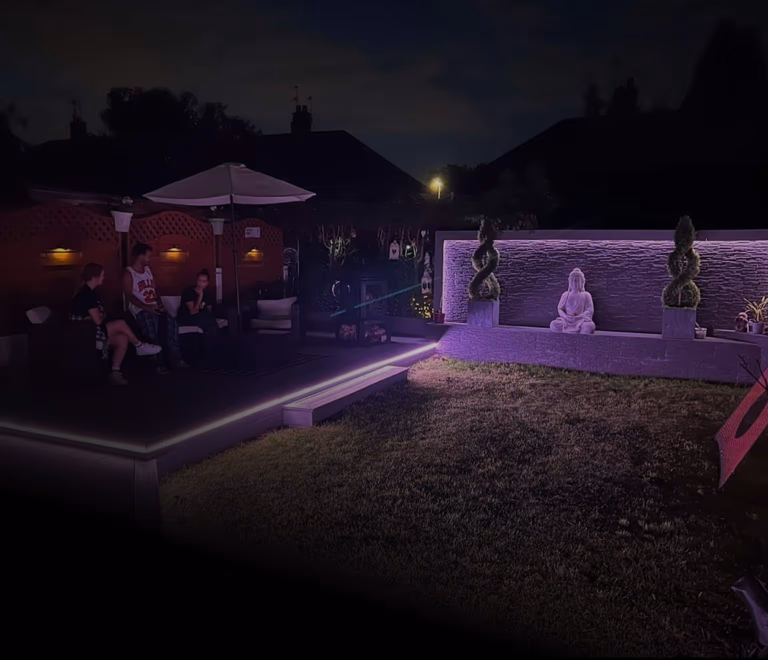 Three people sitting on a garden deck with purple LED lighting at night, facing a Buddha statue and sculpted topiary under soft ambient lights.