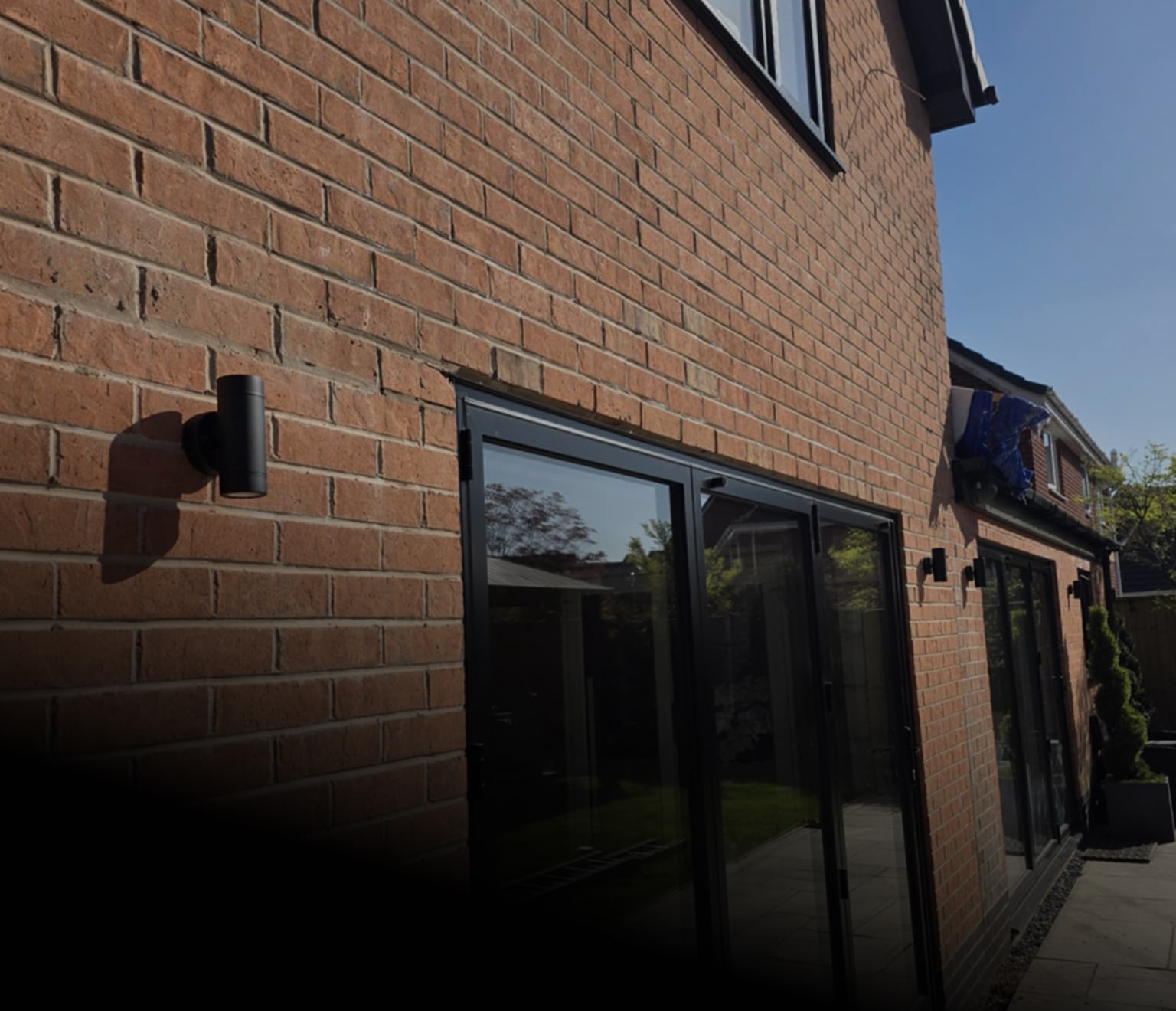 Exterior side view of a brick house with black-framed glass sliding doors and cylindrical outdoor wall lights mounted on the wall.