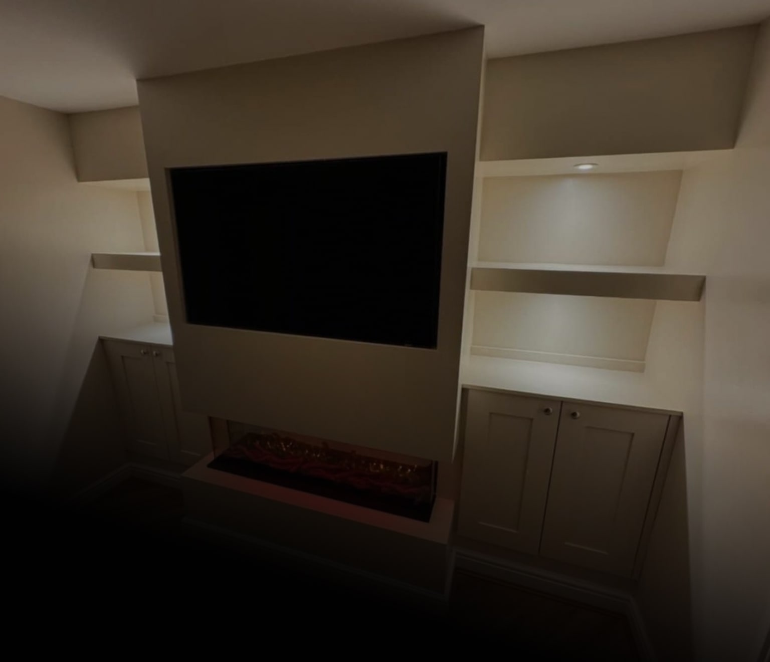 Built-in wall unit with a large blank TV screen above an electric fireplace, flanked by white shelves and cabinets with recessed lighting.