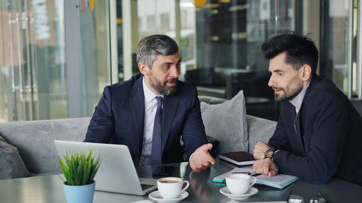 Two men in suits talking at a table with a laptop, notebooks, and coffee cups
