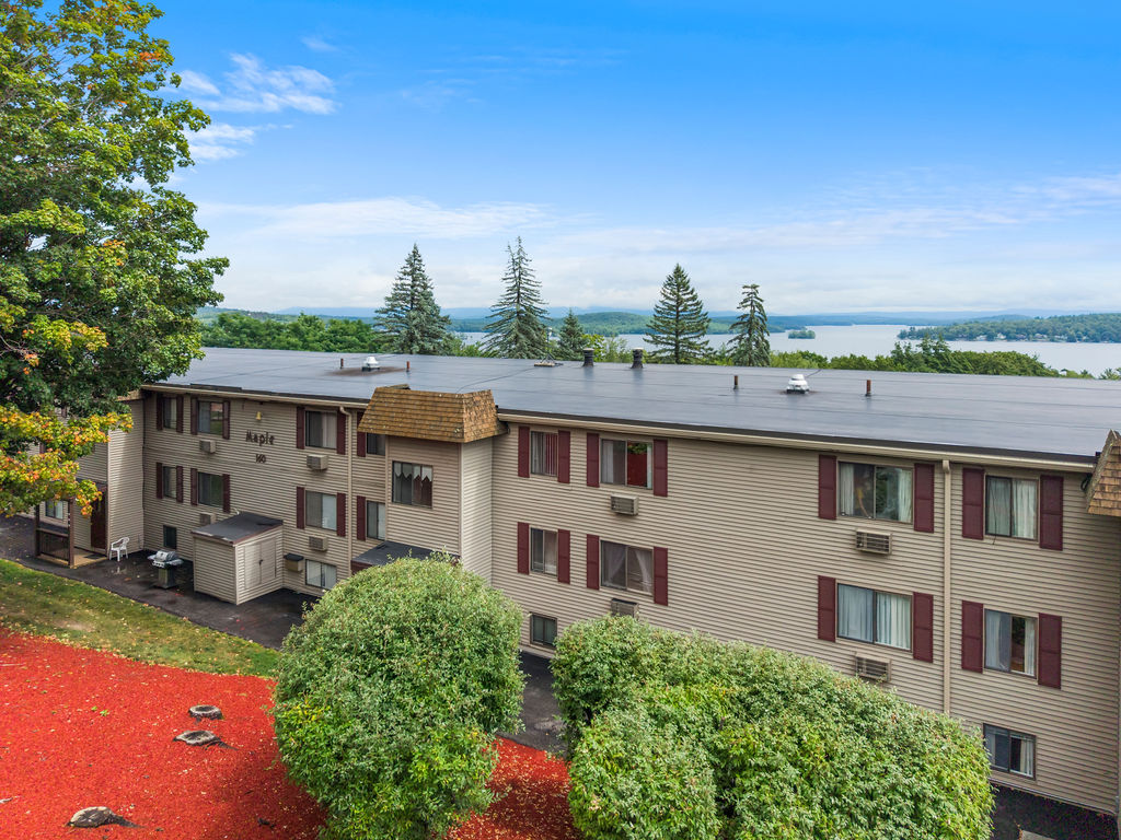 Three-story beige apartment building named Maple with maroon shutters surrounded by green bushes and trees, overlooking a large lake with islands under a blue sky with scattered clouds.