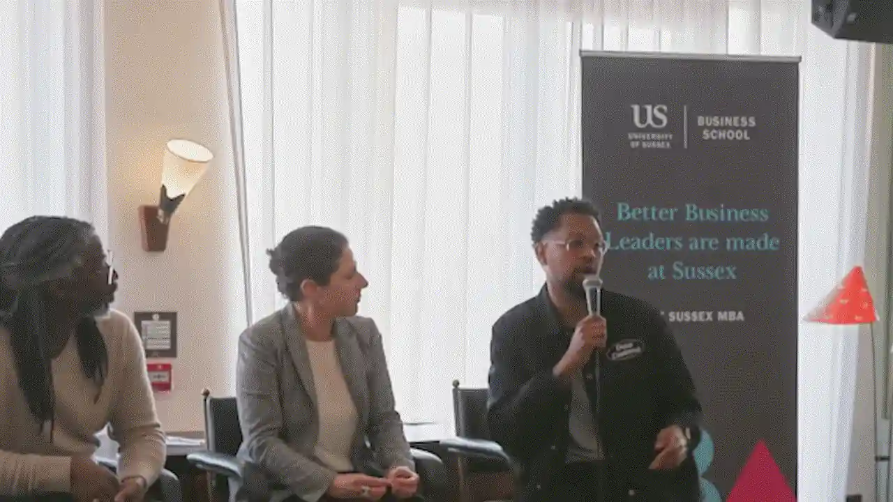 A panellist with a microphone speaking beside a University of Sussex Business School banner.